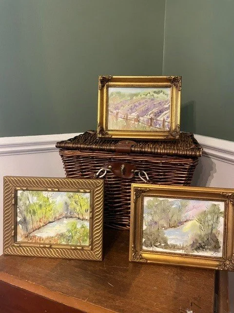 Three small landscape paintings with gold and silver frames, resting on a wooden surface and a wicker basket, against a green wall with white trim.