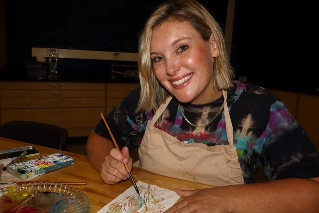 A woman smiling and painting at a wooden table, wearing an apron and a colorful tie-dye shirt, in an indoor setting.