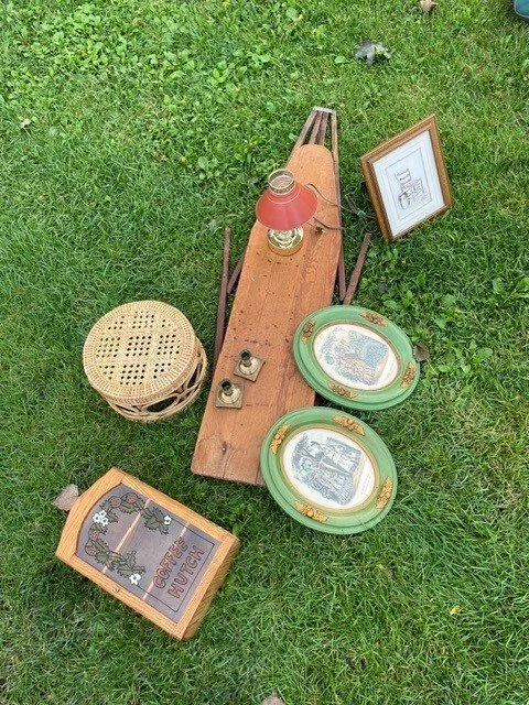 A collection of vintage home decor items on a grassy lawn, including a wooden sled, framed artwork, a lamp, decorative plates, a wicker stool, and a wooden framed plaque.