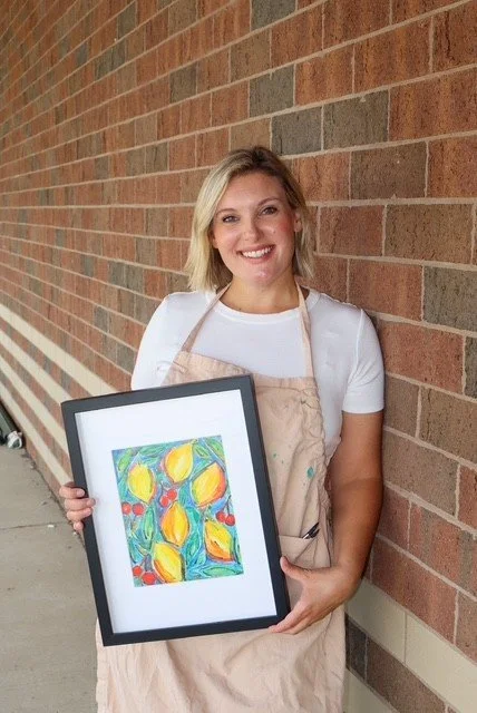 The artist is smiling and holding a framed colorful abstract painting, standing against a brick wall.