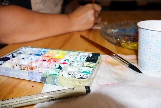 Painting supplies on a table, including watercolor paints, brushes, a mug, and a paper towel.