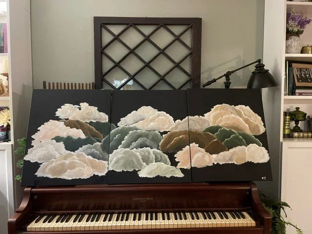 Three-panel painting of clouds on top of a dark brown piano, with a black wall-mounted lamp, a mirror, and shelves in the background.