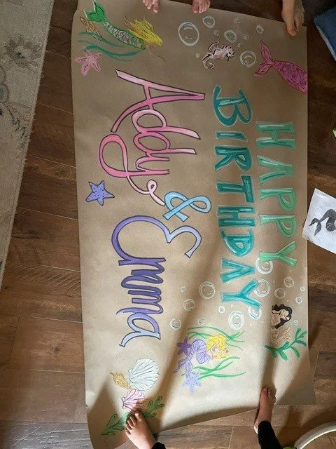 Colorful birthday banner with underwater theme featuring mermaids, sea creatures, and decorative bubbles. The banner has a message that reads, "HAPPY BIRTHDAY Addy & Emma."
