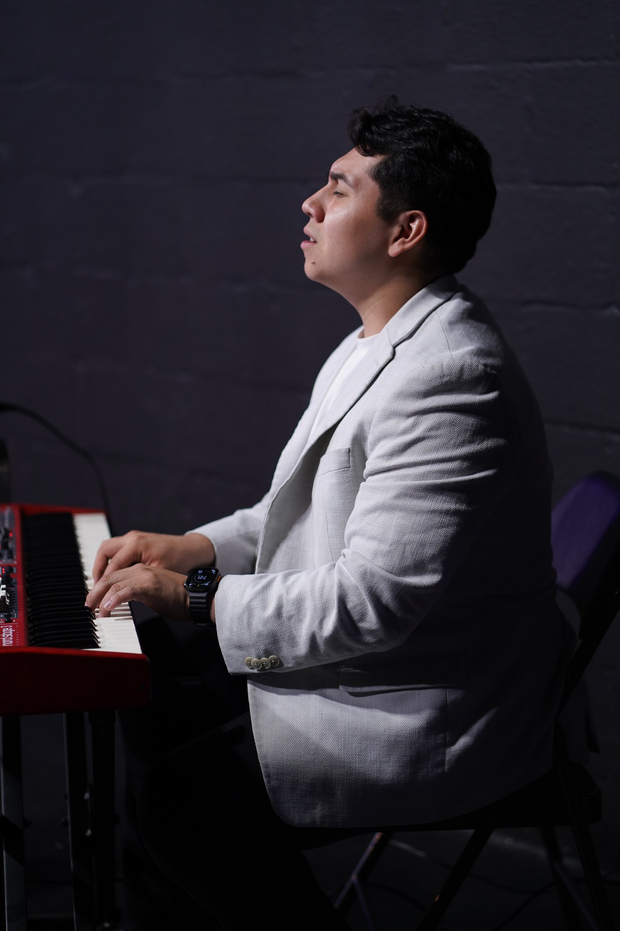 A man in a light-colored blazer playing worship music a red and black digital keyboard with eyes closed, seated at at Acts Church Hollywood.