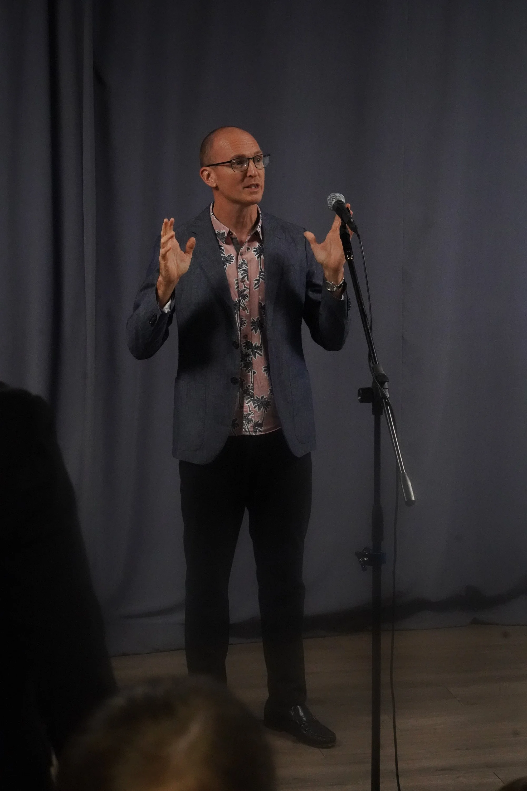A man with glasses, dressed in a blazer and patterned shirt, standing in front of a dark grey wall, speaking into a microphone at Acts Church Hollywood.