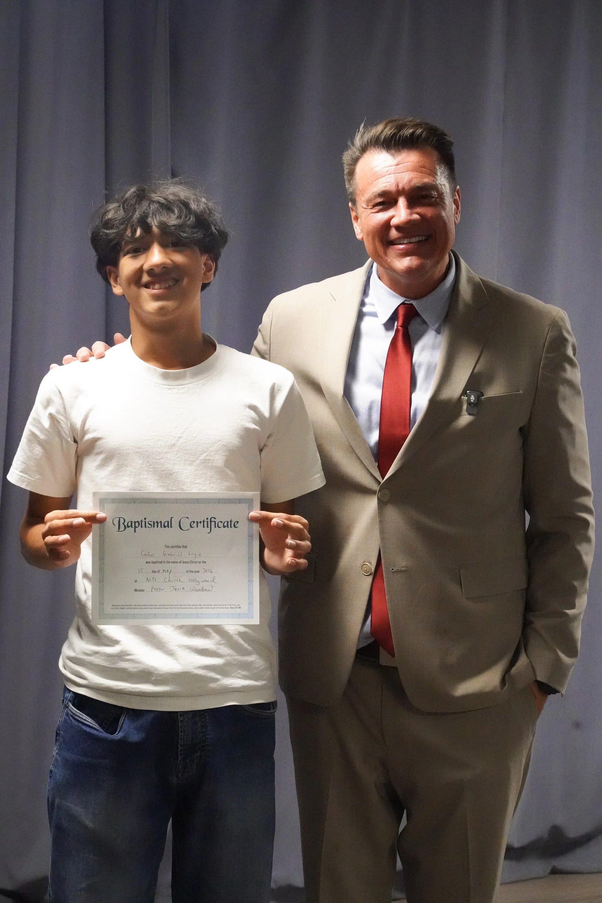 A young boy with dark curly hair holding a baptismal certificate standing next to a man in a tan suit and red tie with dark hair, smiling in front of a gray curtain.