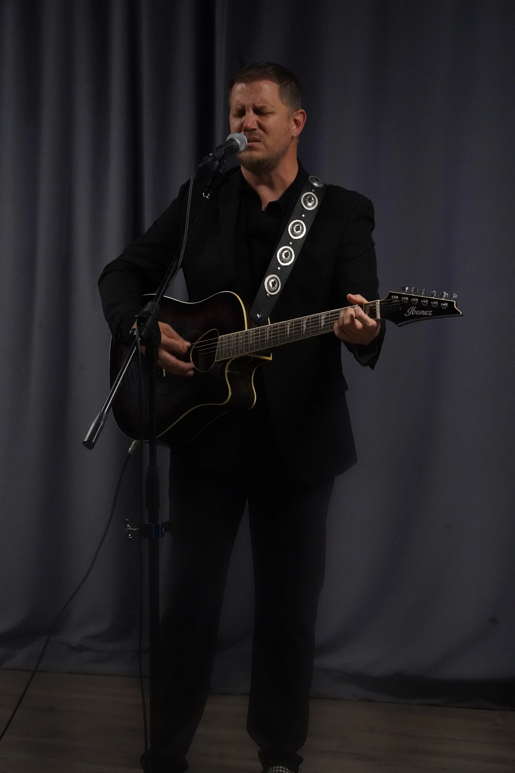 A Christian man dressed in black is singing into a microphone while playing worship music with an acoustic guitar at Acts Church Hollywood.