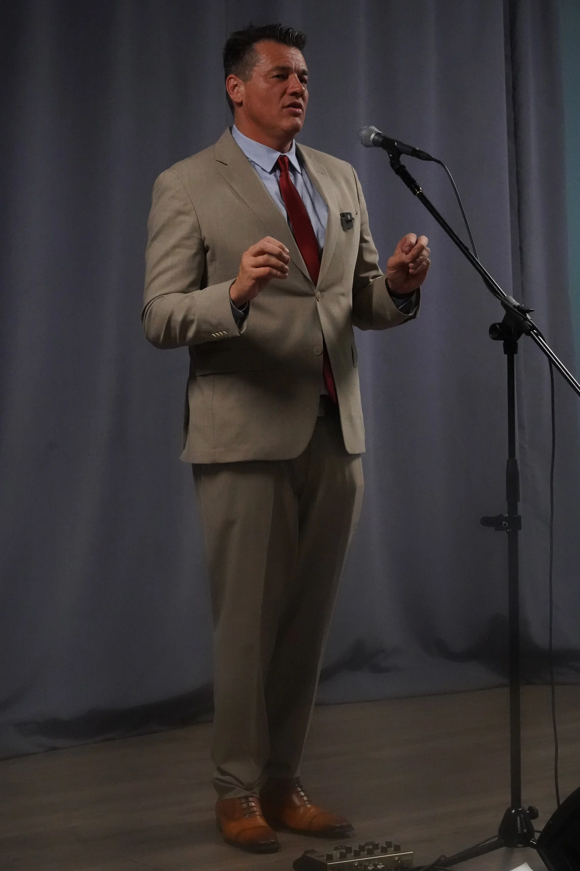 Man in a beige suit, light blue shirt, red tie, and brown shoes standing and speaking into a microphone on a stand, with a gray curtain backdrop.