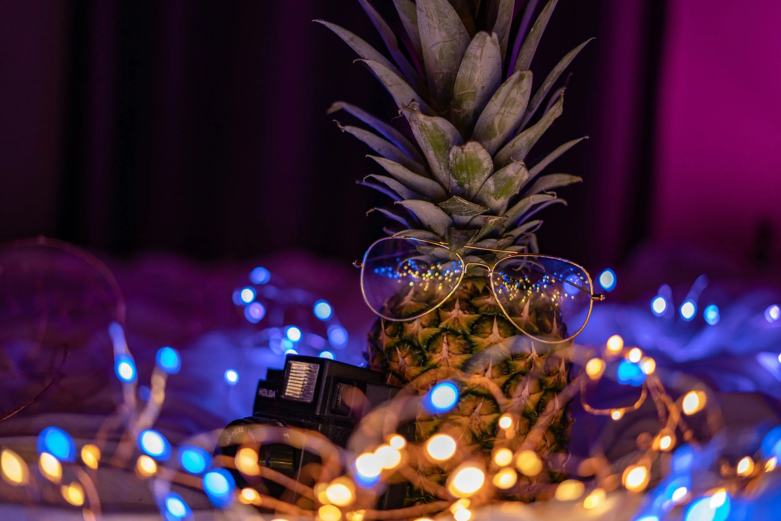 A pineapple with glasses resting on its leaves, surrounded by colorful string lights with blue, yellow, and purple hues.