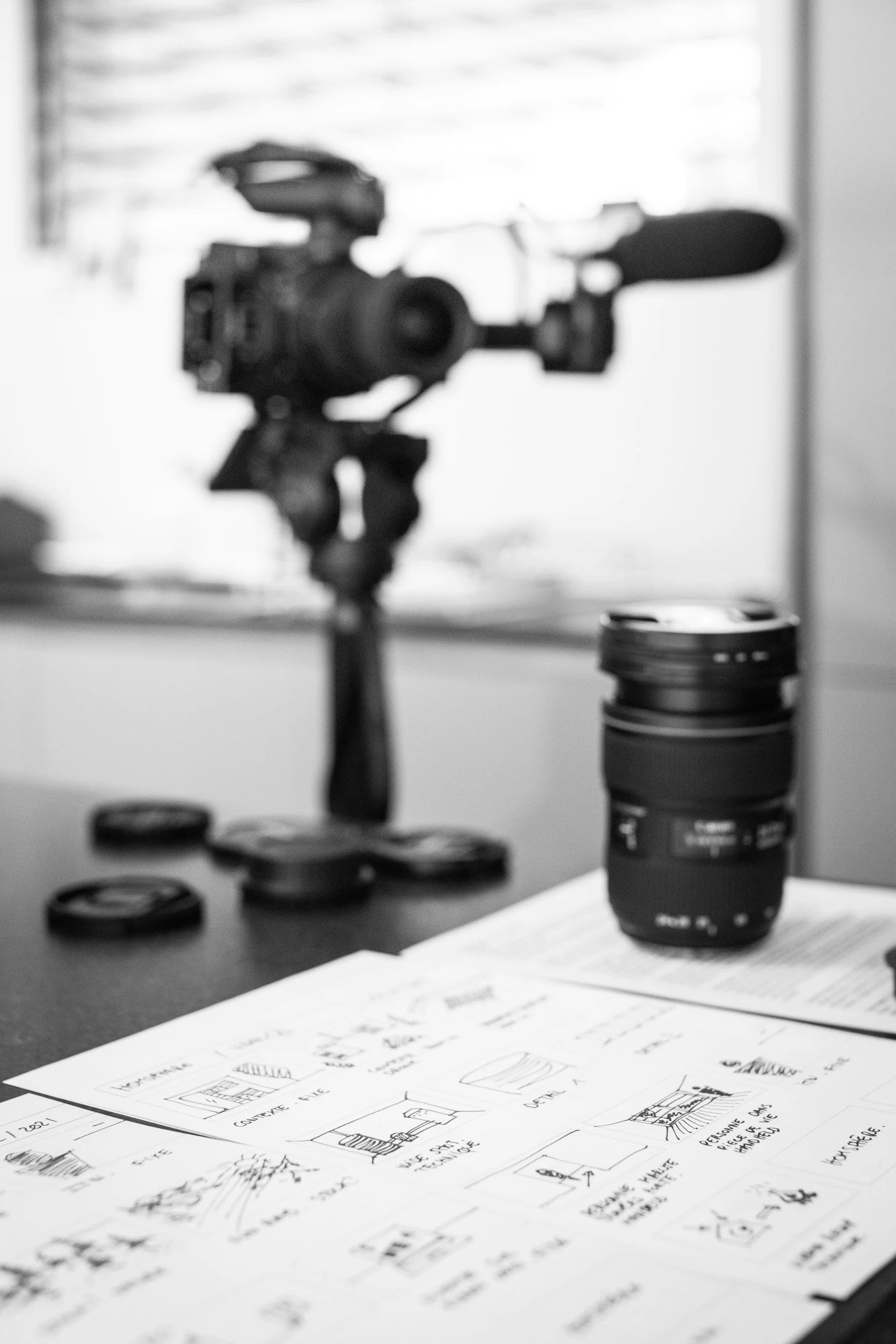 Black-and-white photograph showing architectural sketches on paper spread across a table, with a camera lens and a camera mounted on a tripod in the background, along with camera lens caps on the table.