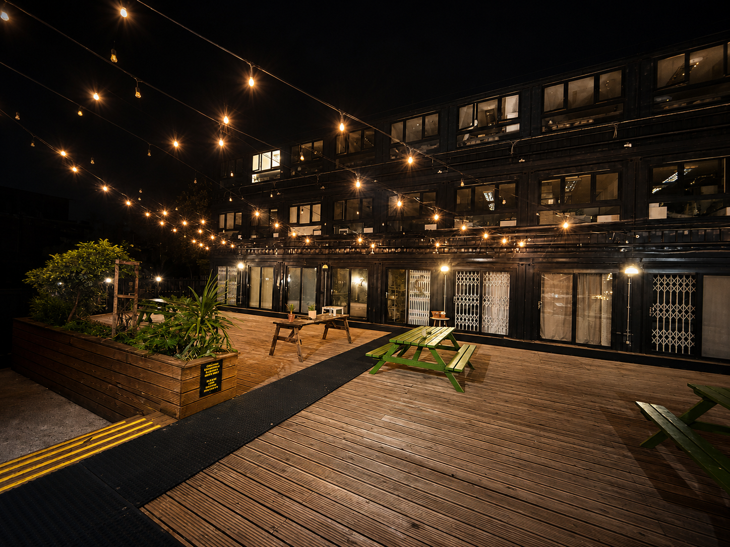 The First Studio Court yard at Night - Night time outdoor patio with string lights, wooden benches, and a green picnic table in front of a modern black building with large glass windows.