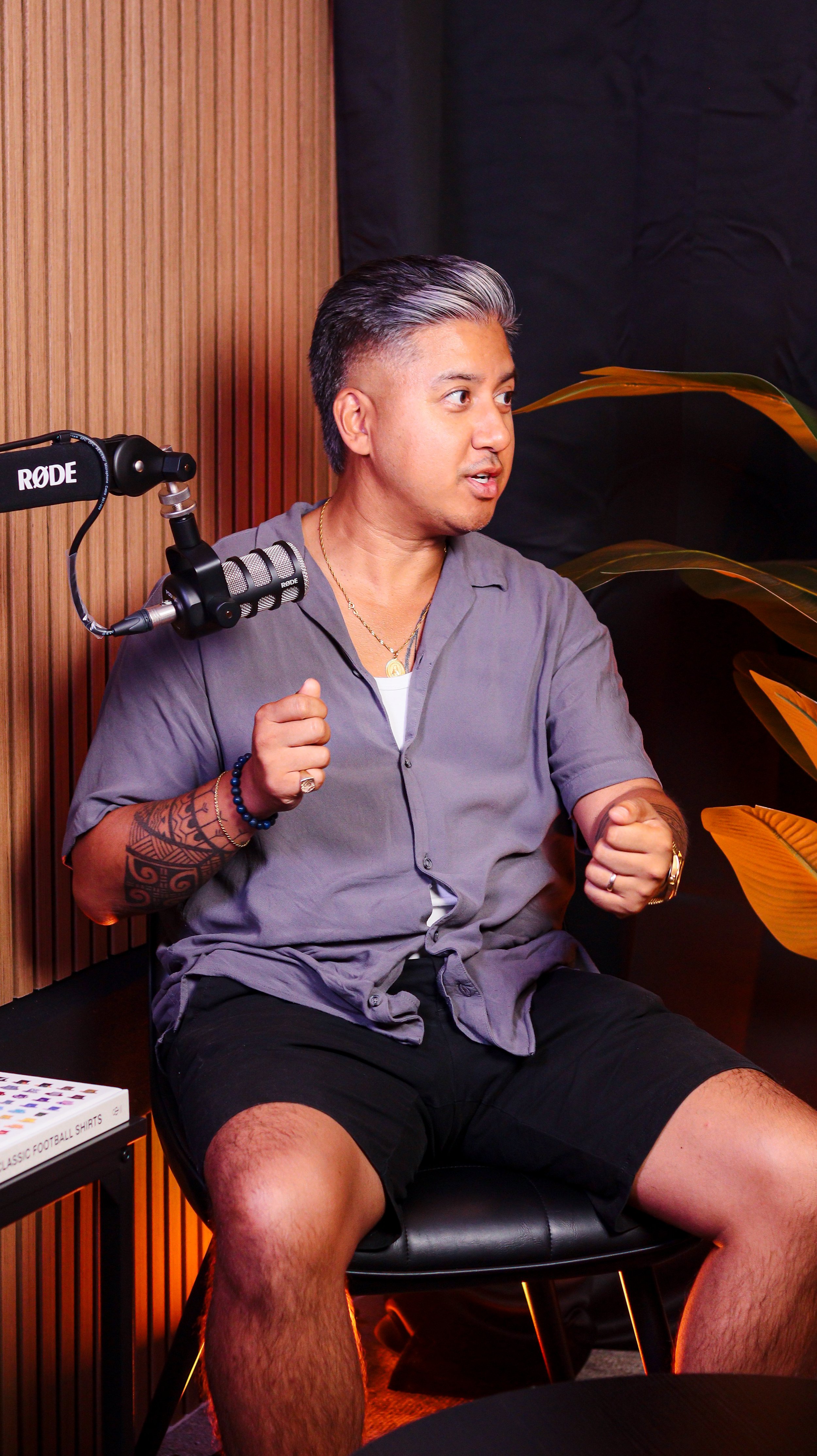 A young man sitting in a chair with a microphone in front of him, gesturing with his hands while speaking. He has short dark hair with gray highlights and is wearing a gray short-sleeved button-up shirt, black shorts, and jewelry. There is a wooden wall and some plants in the background.