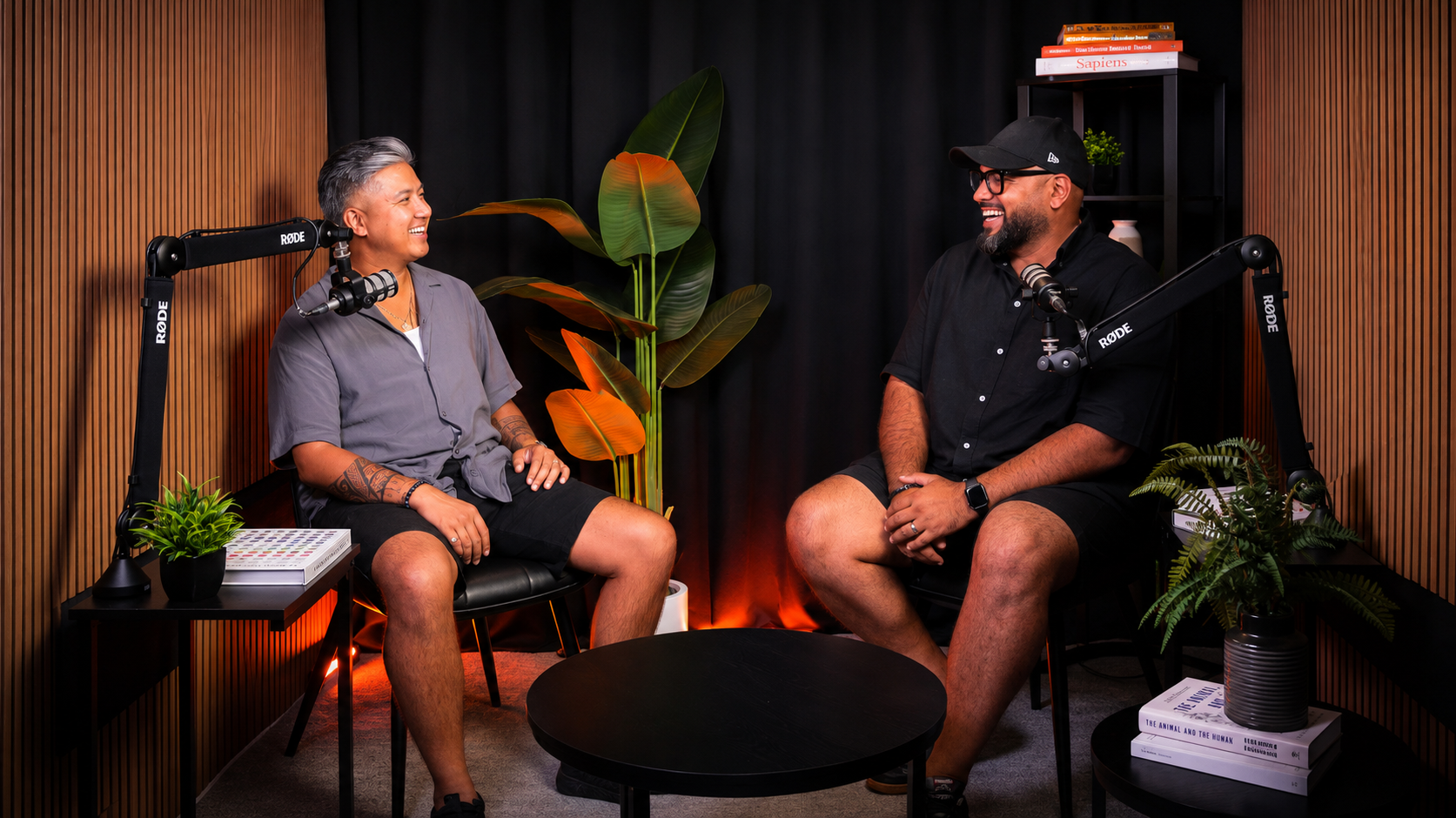 Two men are sitting in a podcast studio, smiling and speaking into microphones with Rode branding. The studio has wood-paneled walls, a large plant with broad leaves, and shelves with books. Small potted plants are on side tables, and a black round table is in the center.