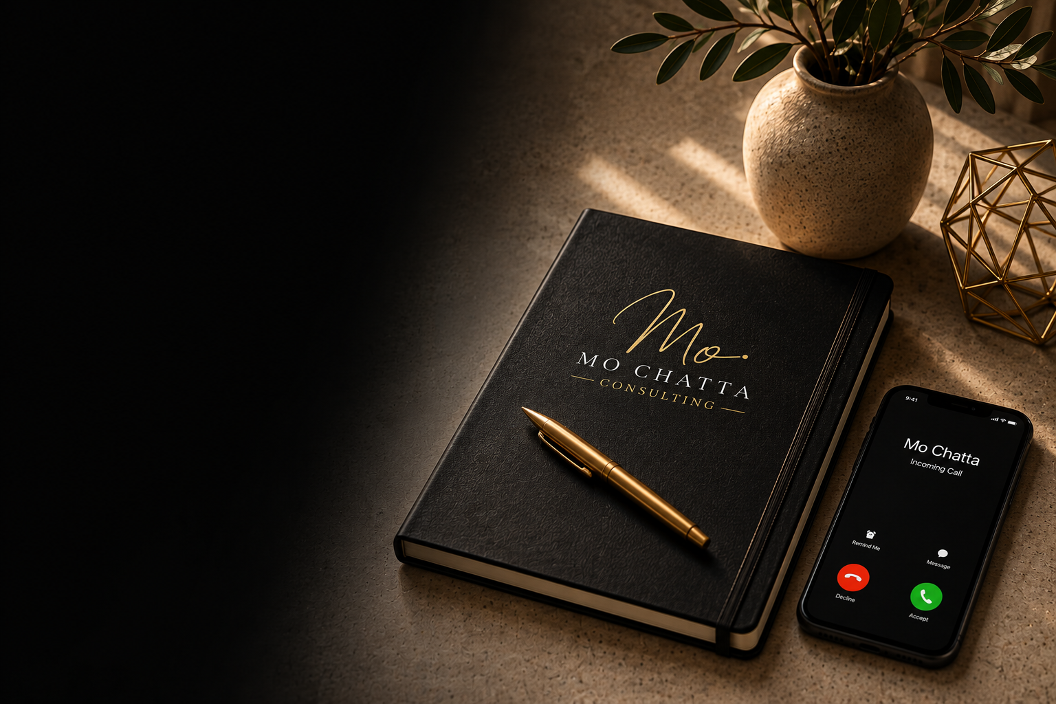 A black notebook with gold text reading 'Mo Chatta Consulting,' a gold pen placed on top, a smartphone displaying an incoming call from Mo Chatta, a beige textured vase with green leaves, and a geometric gold decorative object, arranged on a tan surface with a soft light