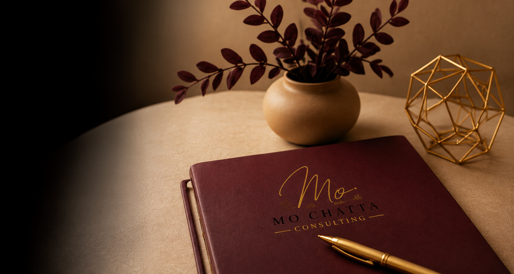 A desk with a burgundy notebook titled "Mo Chatta Consulting," a gold pen, a beige vase with purple leaves, and a gold geometric sculpture.