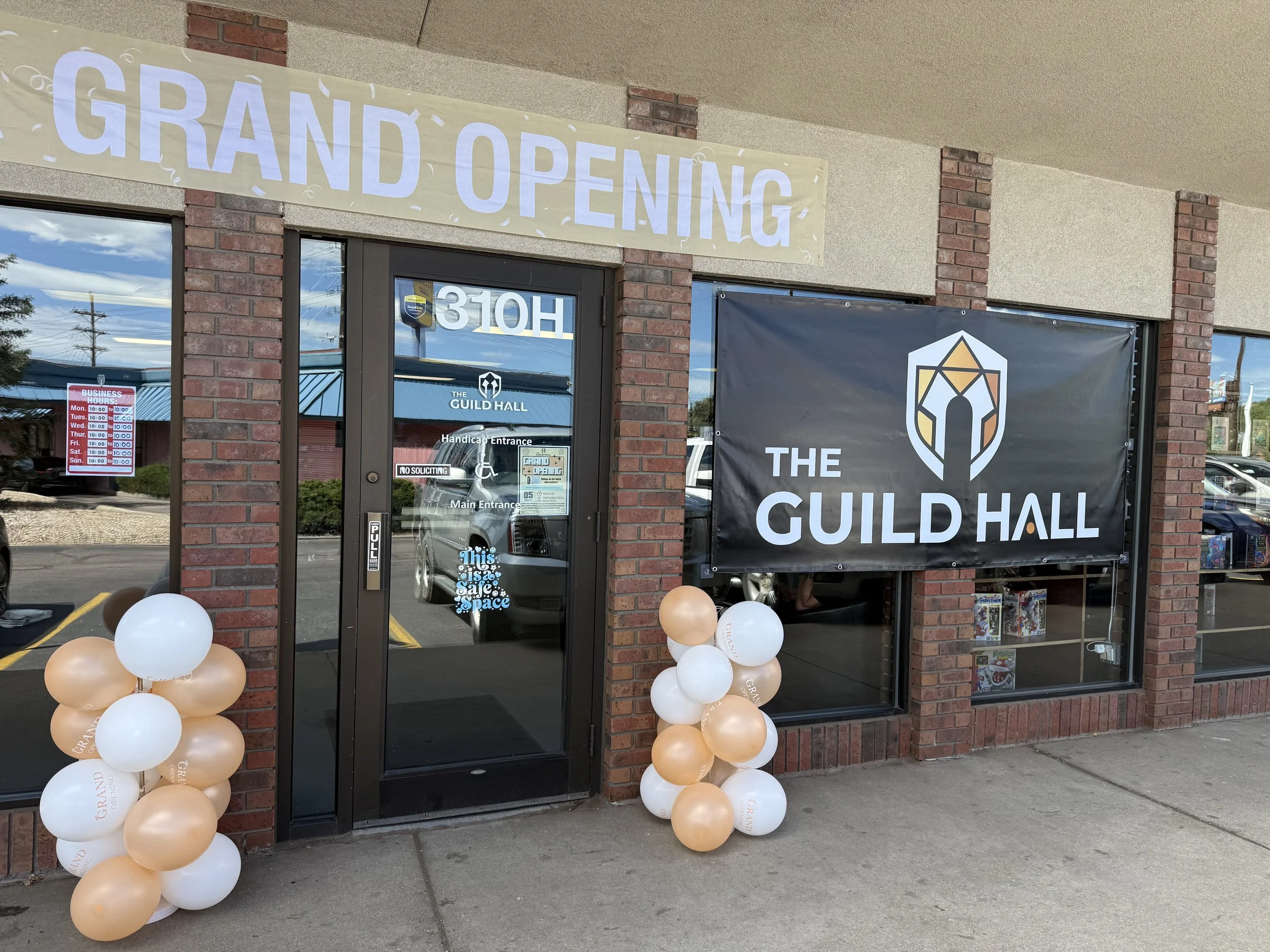 Store entrance decorated with a large beige banner that reads 'Grand Opening', and a large black banner with a logo that says 'The Guild Hall'. Balloons in white and beige are placed around the entrance.