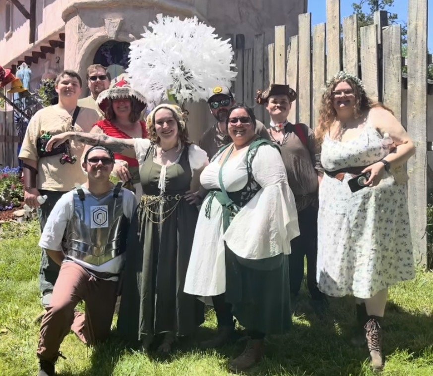 Group of nine people dressed in medieval and fantasy costumes, posing outdoors on a sunny day with a wooden fence and a pink building in the background.