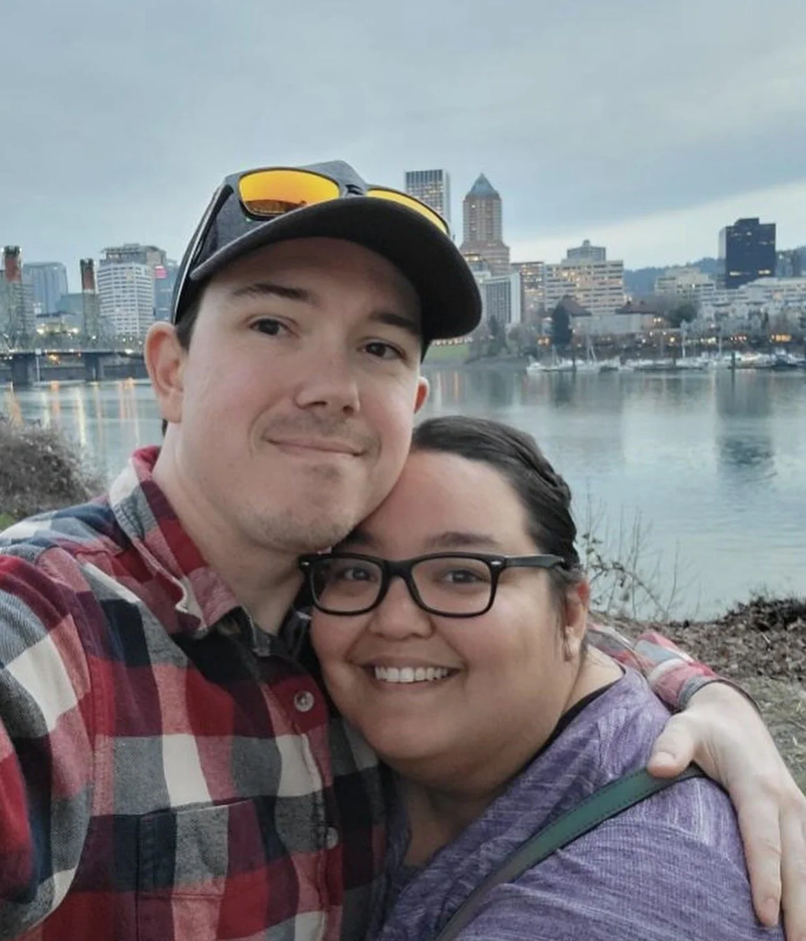 Co-owner of The Guild Hall smiling and hugging his girlfriend by a waterfront with a city skyline in the background during evening or dusk.