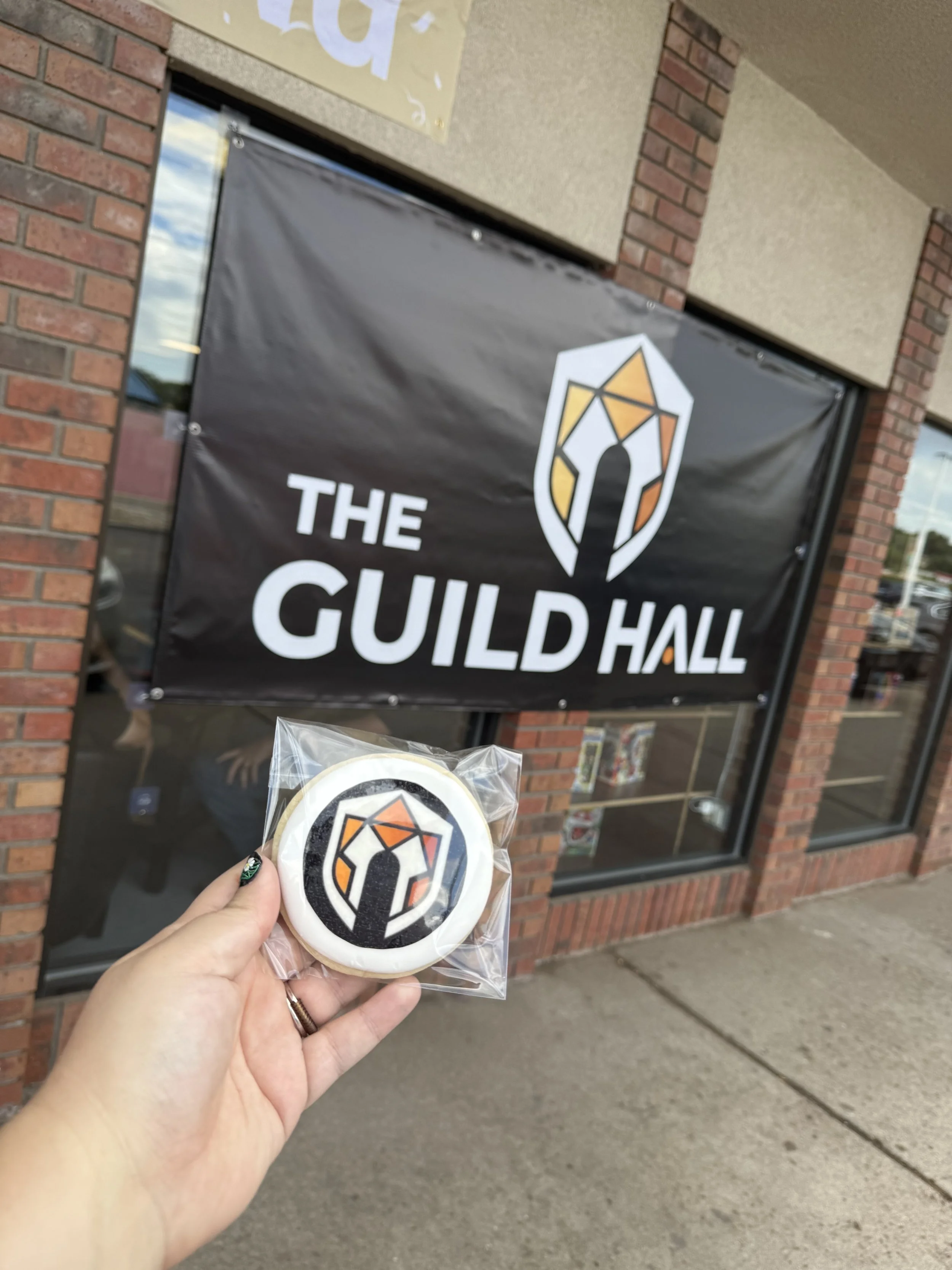 A person holding a packaged cookie with a logo of a stylized shield on it, in front of a sign for 'The Guild Hall' outside a brick building with large glass windows.