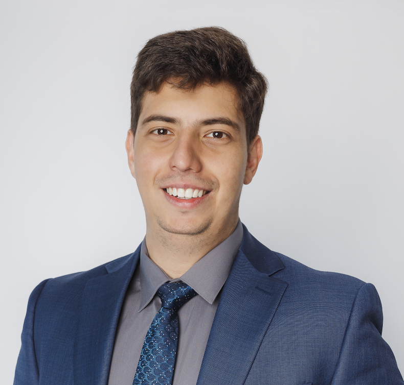 A young man with brown hair, wearing a blue suit, gray shirt, and blue patterned tie, smiling at the camera against a plain white background.