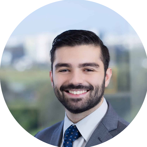 Professional headshot of a smiling man with dark hair and a beard, wearing a gray suit and blue tie, against a blurred outdoor cityscape background.