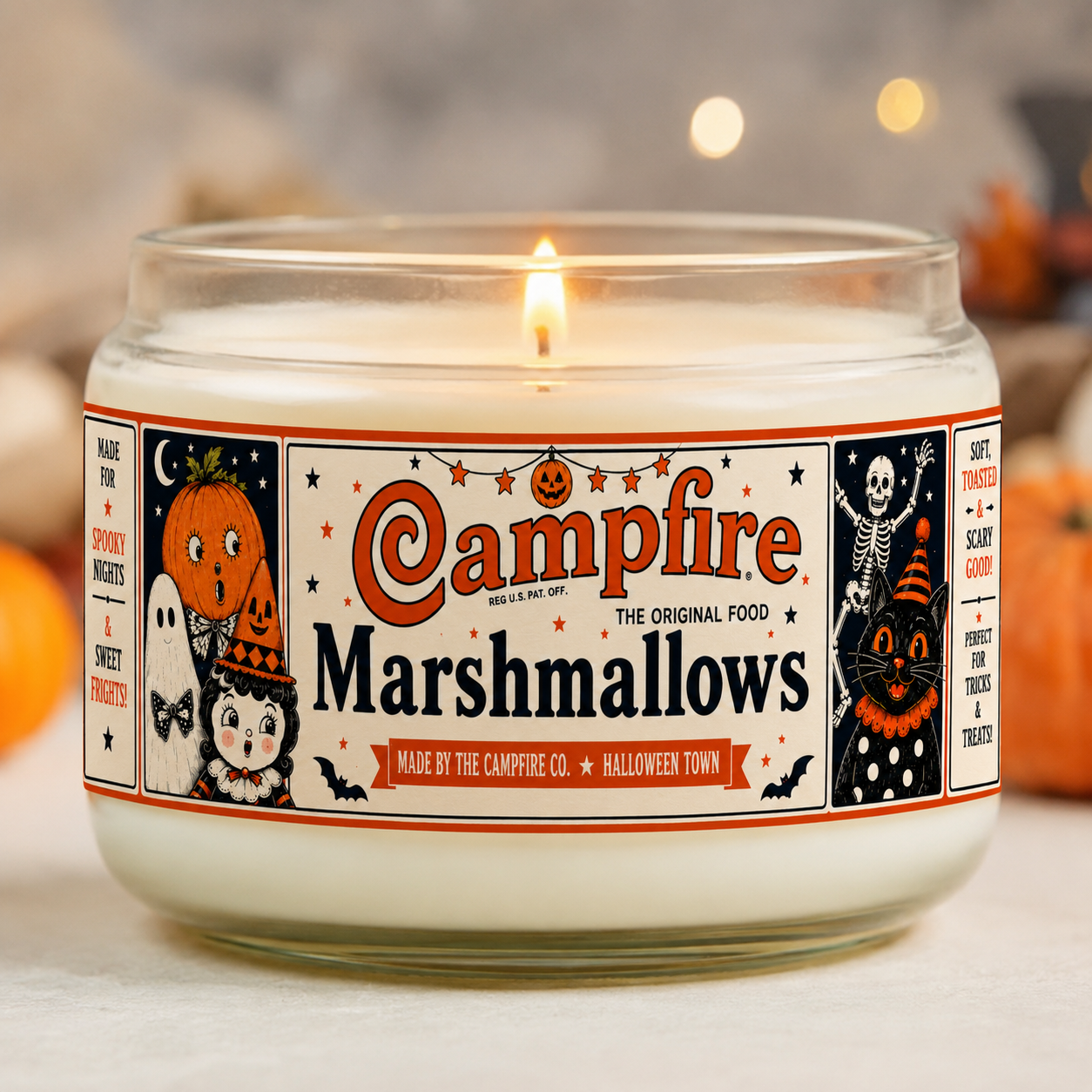 A lit candle with a Halloween-themed label reading 'Campfire Marshmallows' and featuring cartoon Halloween characters and decorations. The background has soft, out-of-focus autumnal decor.