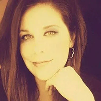 A woman with long brown hair and light-colored eyes smiling, resting her chin on her hand, with a dark background.