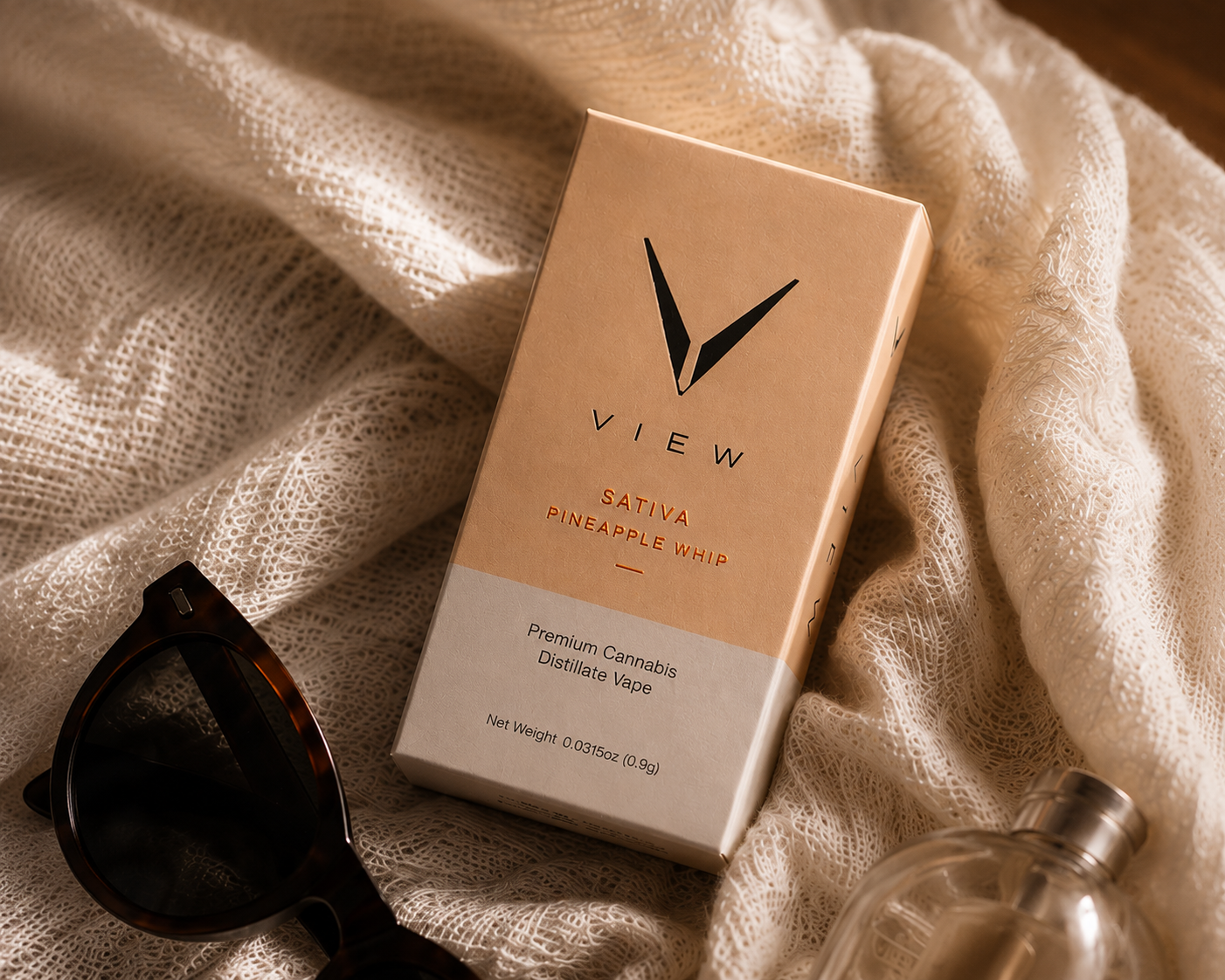 A box of Vew Sativa Pineapple Whip cannabis vape lies on cream-colored lace fabric, with a pair of sunglasses and a bottle nearby.