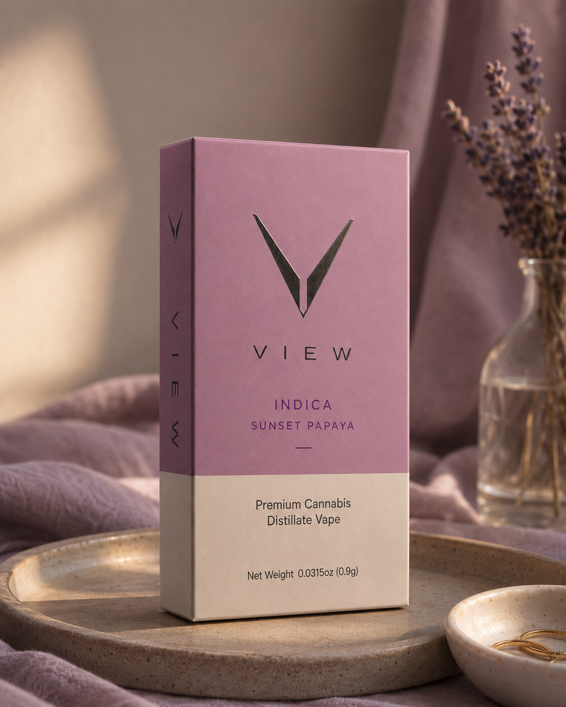 A pink and cream box of VIEW Indica Sunset Papaya cannabis vape with black text, placed on a wooden tray with a purple cloth and a glass vase with lavender flowers in the background