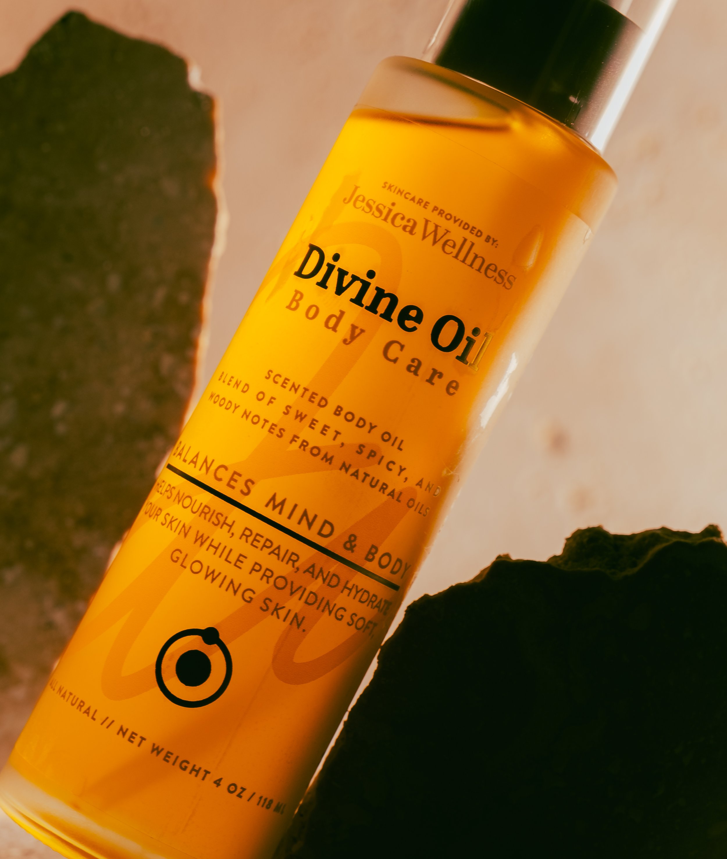 Close-up of a bottle of Jessica Wellness Divine Oil Body Care surrounded by dark green leaves.