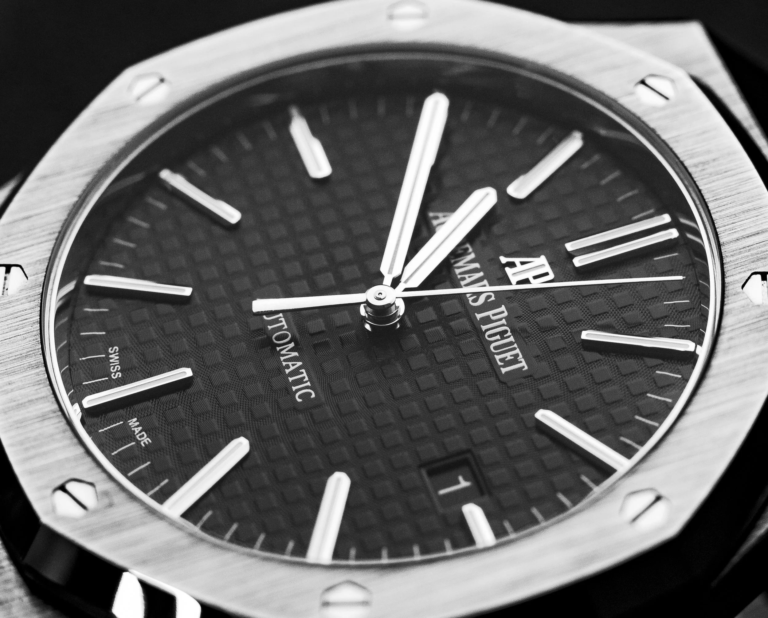 Close-up of a luxury watch with a black textured dial, silver hour markers, and hands, featuring the brand name 'Audemars Piguet' and a date window. The watch has a metal case and screws on the bezel.