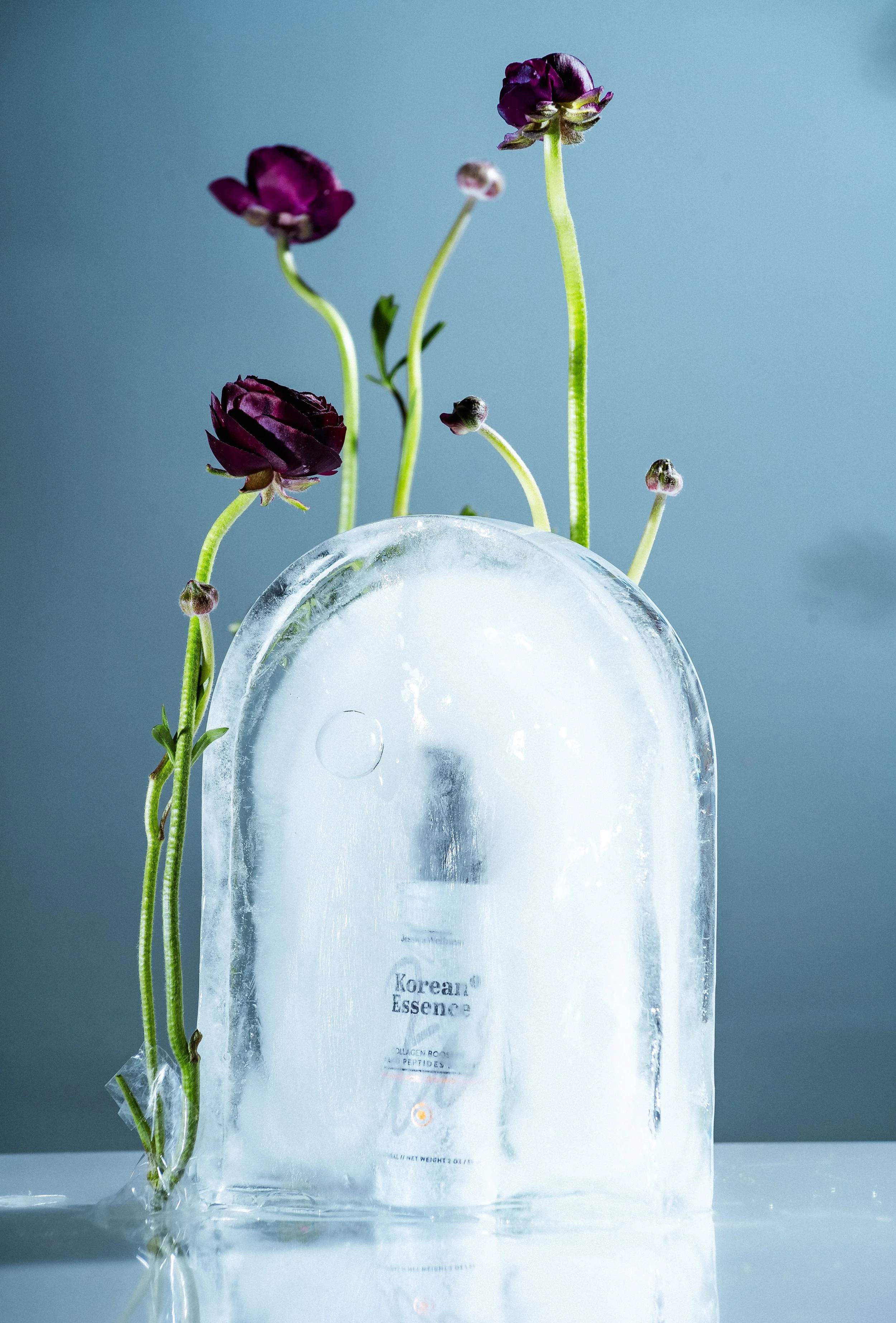 A clear ice block with purple flowers and green stems emerging from the top, placed on a reflective surface with a light blue background.