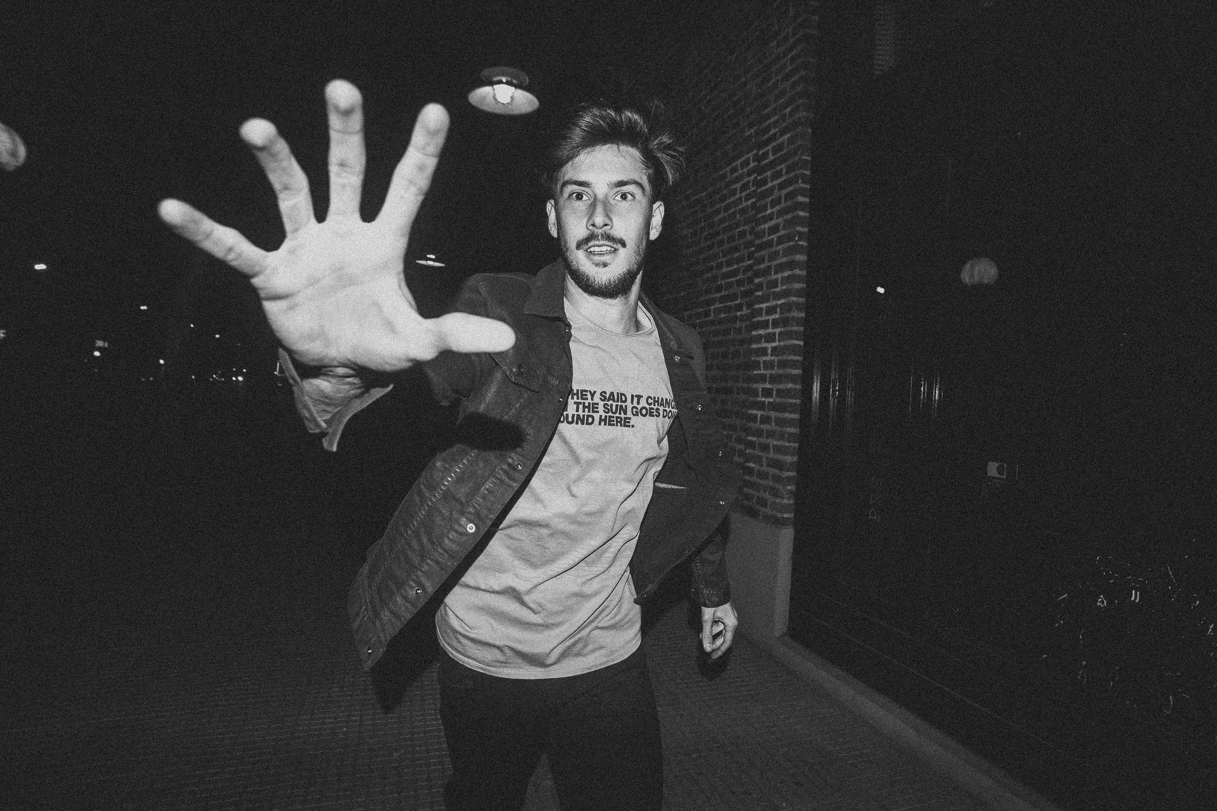 A young man with a beard and styled hair reaching out toward the camera with his hand, standing outdoors at night near a brick wall and a streetlamp, wearing a casual hoodie and denim jacket.