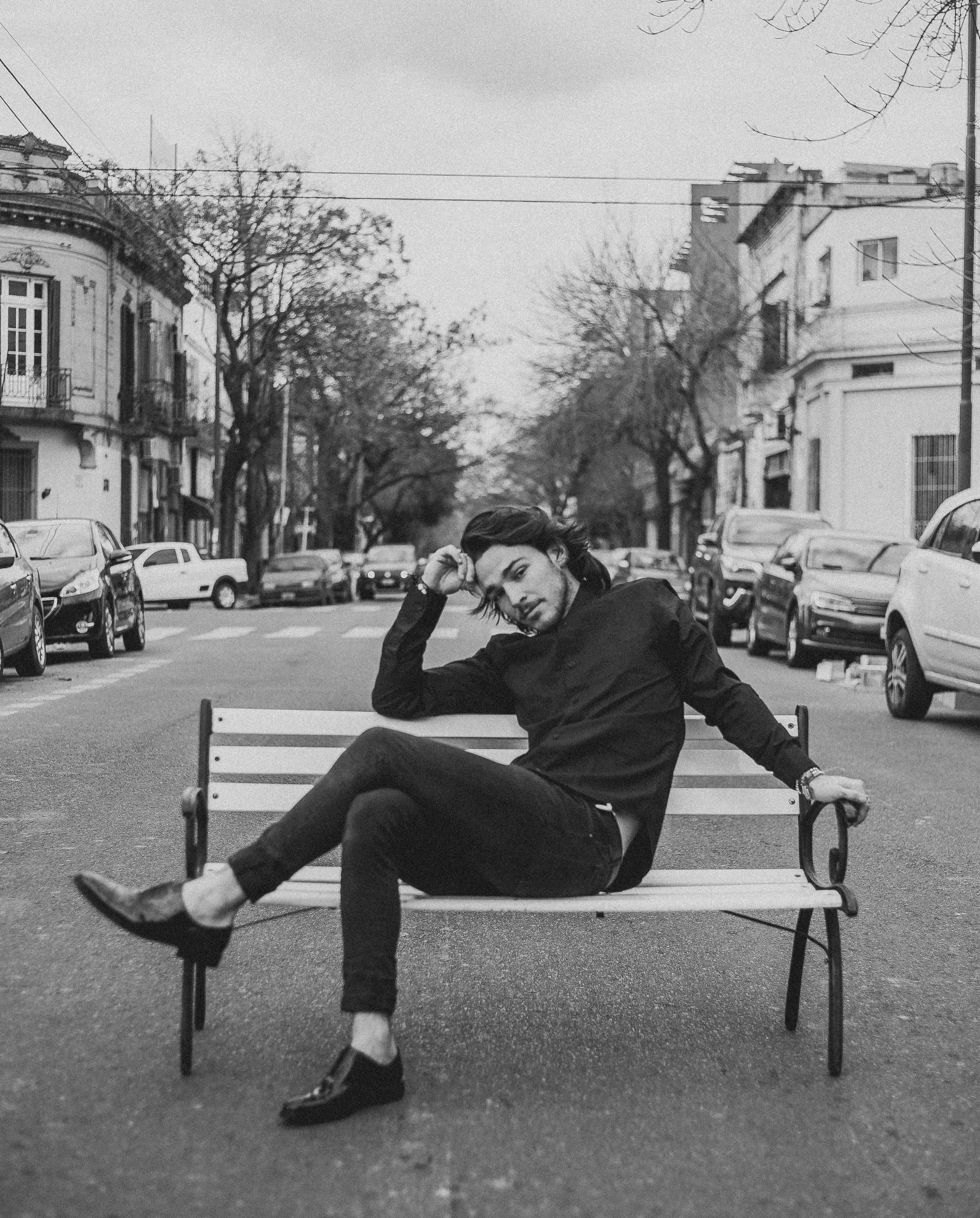 A woman sitting on a park bench in an urban area during winter or early spring. She is dressed in black, with short hair, and is adjusting her hair or scratching her head while looking at the camera.