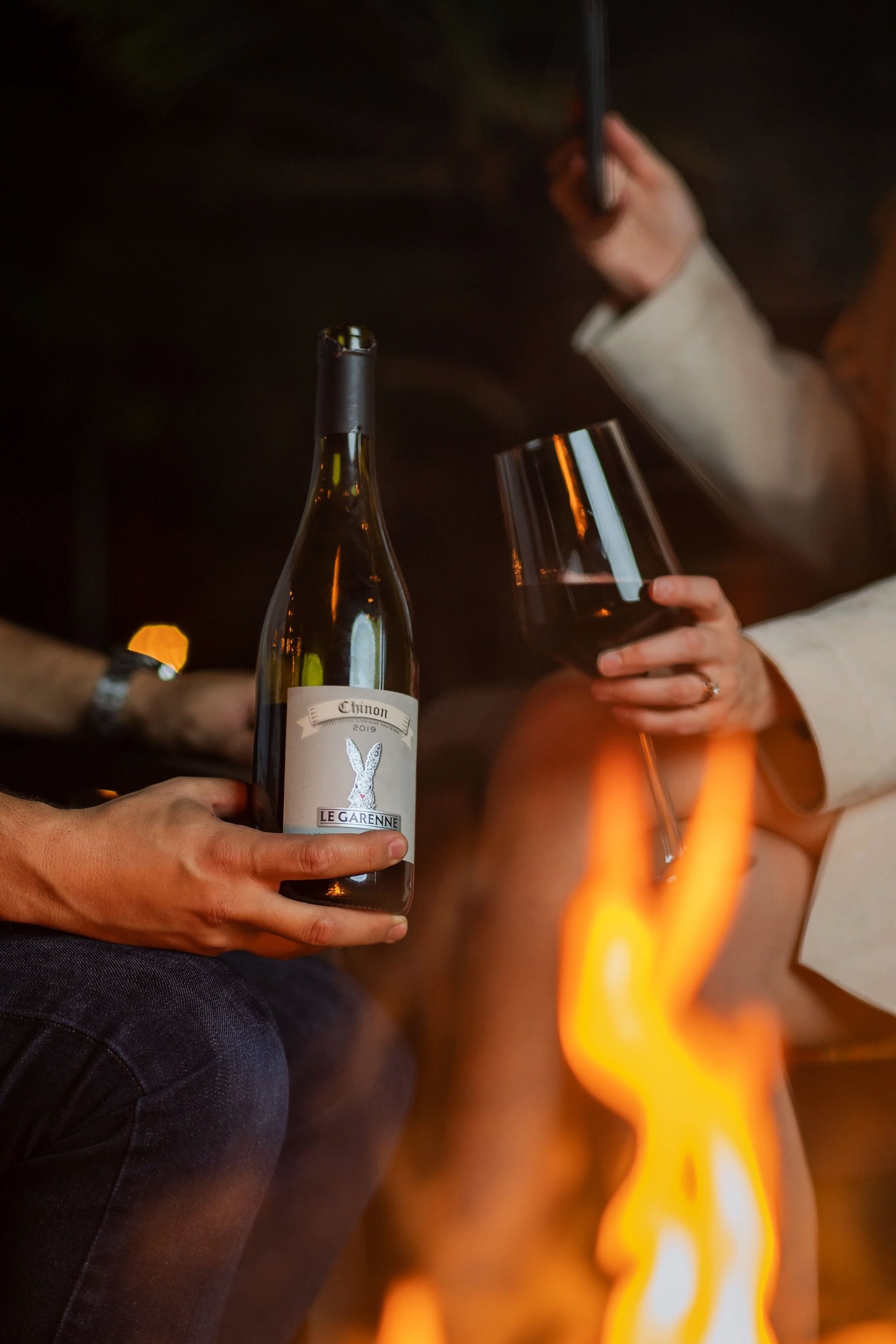 Person holding a glass of red wine, with a bottle of wine labeled "Chinon 2019" and a hand in the background holding a smartphone. There is a flame in the foreground.