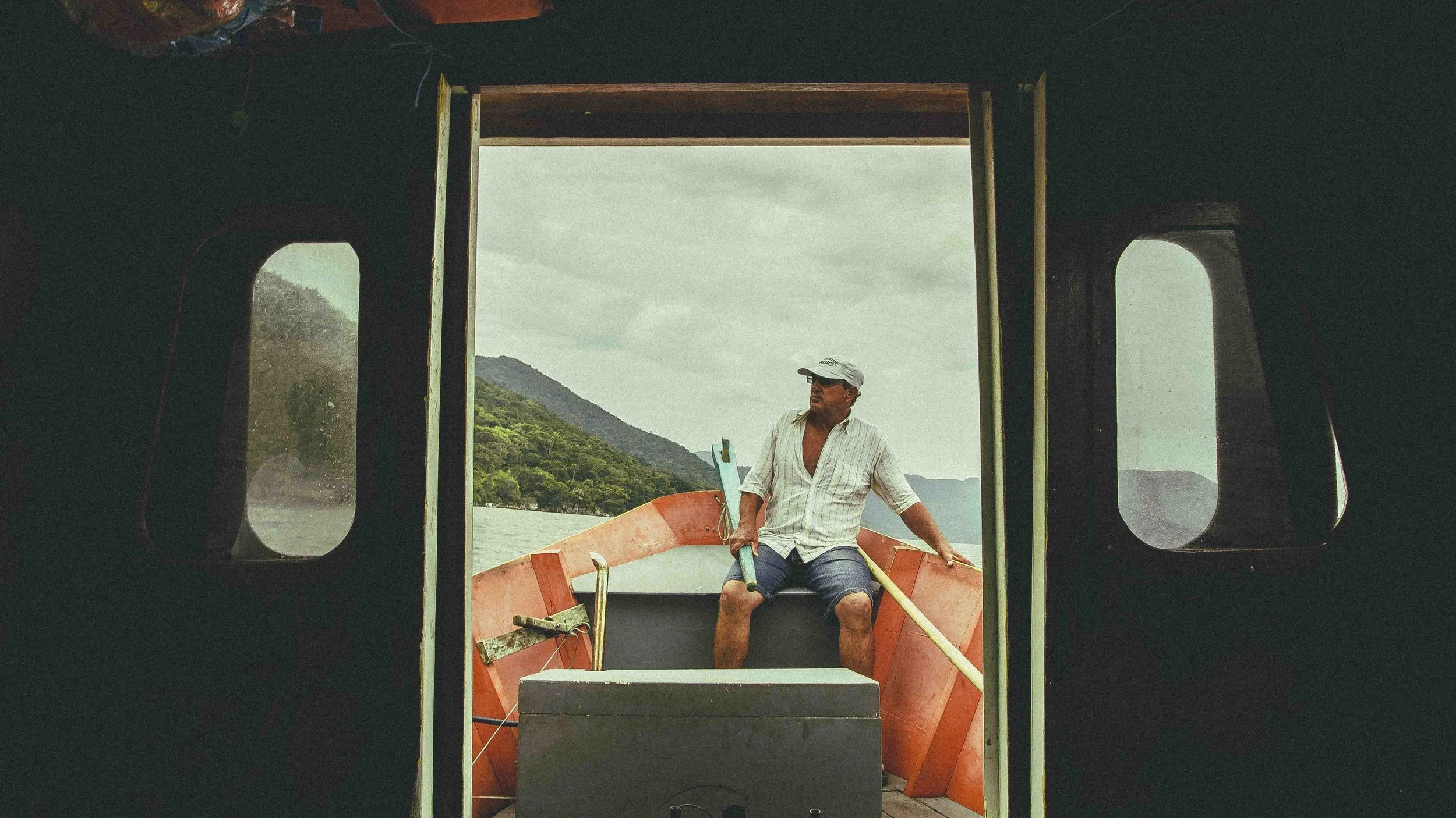 View through a boat window showing a man sitting on the boat with a boat paddle, with water, trees, and mountains visible in the background.