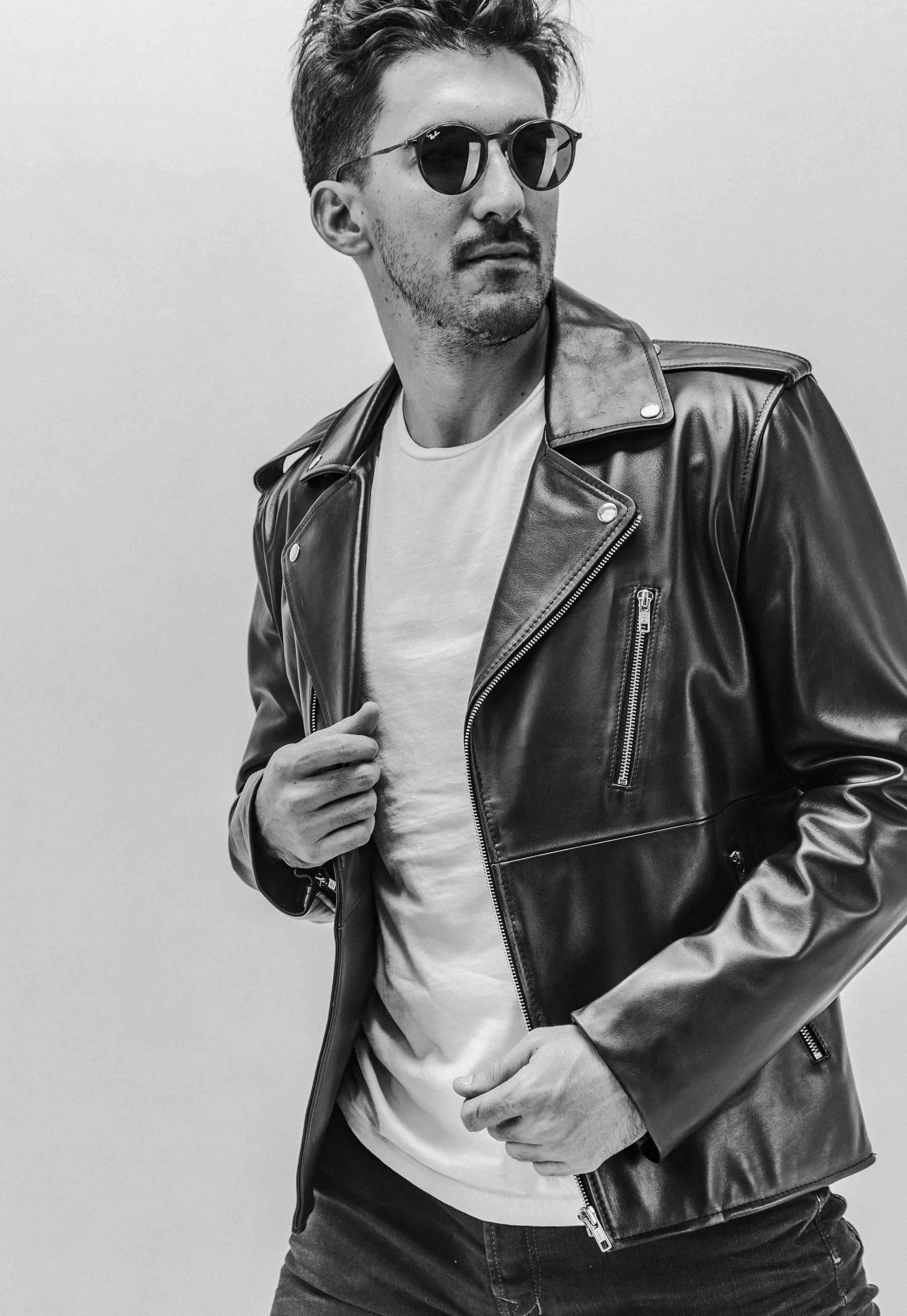 Black and white photo of a man wearing sunglasses, a leather jacket, and a white t-shirt, posing with one hand on his chest and the other near his waist against a plain background.