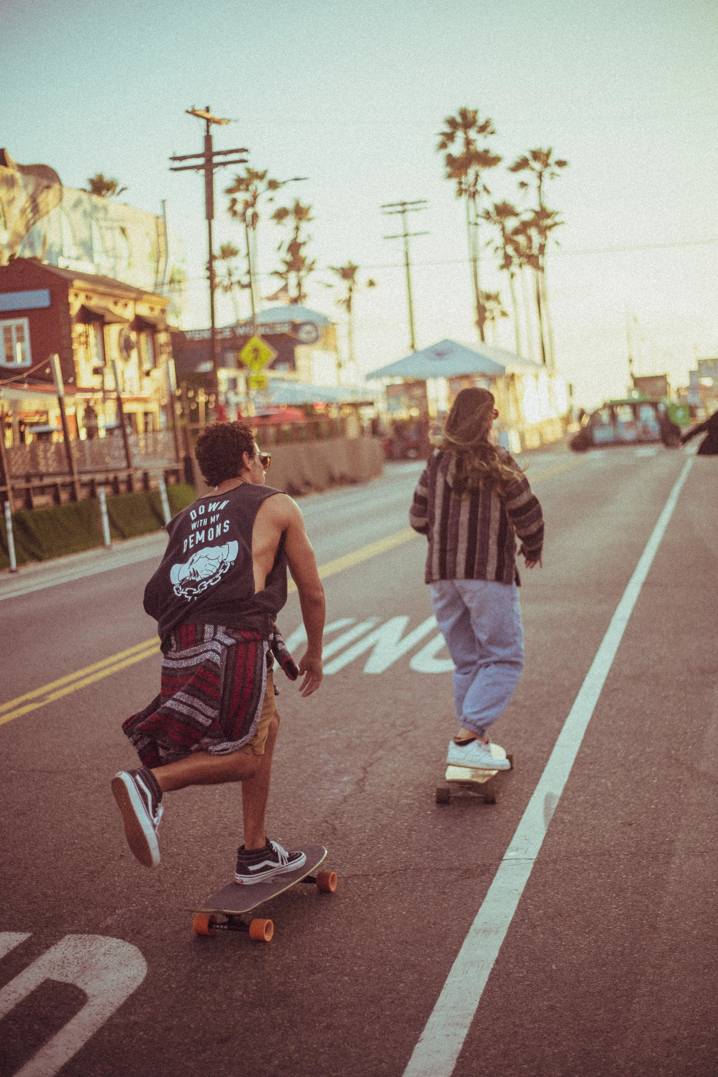 Two young people, one skateboarding and the other walking, along a street with colorful buildings and tall palm trees in the background, during sunset.