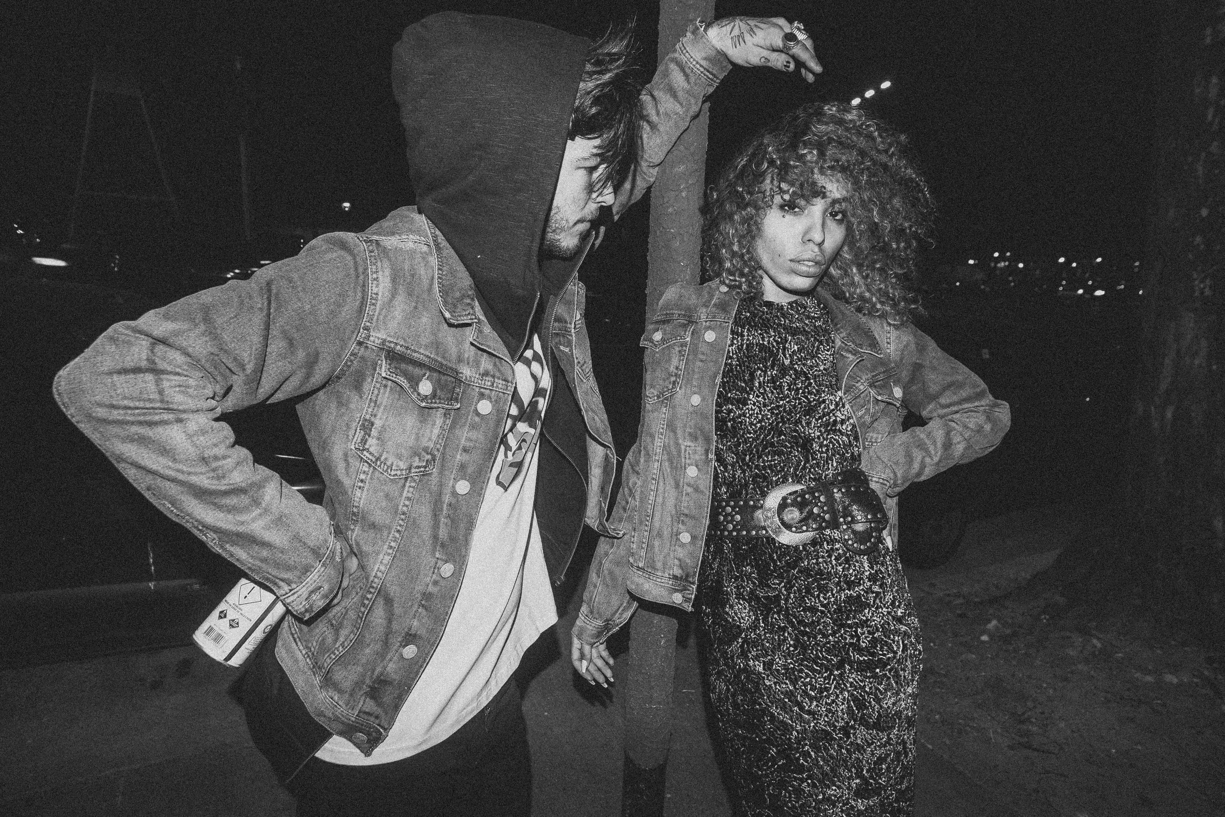 Two people resembling a man and woman are standing outdoors at night. The man on the left has long hair, is wearing a hoodie and a denim jacket, and is holding a drink. The woman on the right has curly hair, is wearing a leopard print dress, a denim 