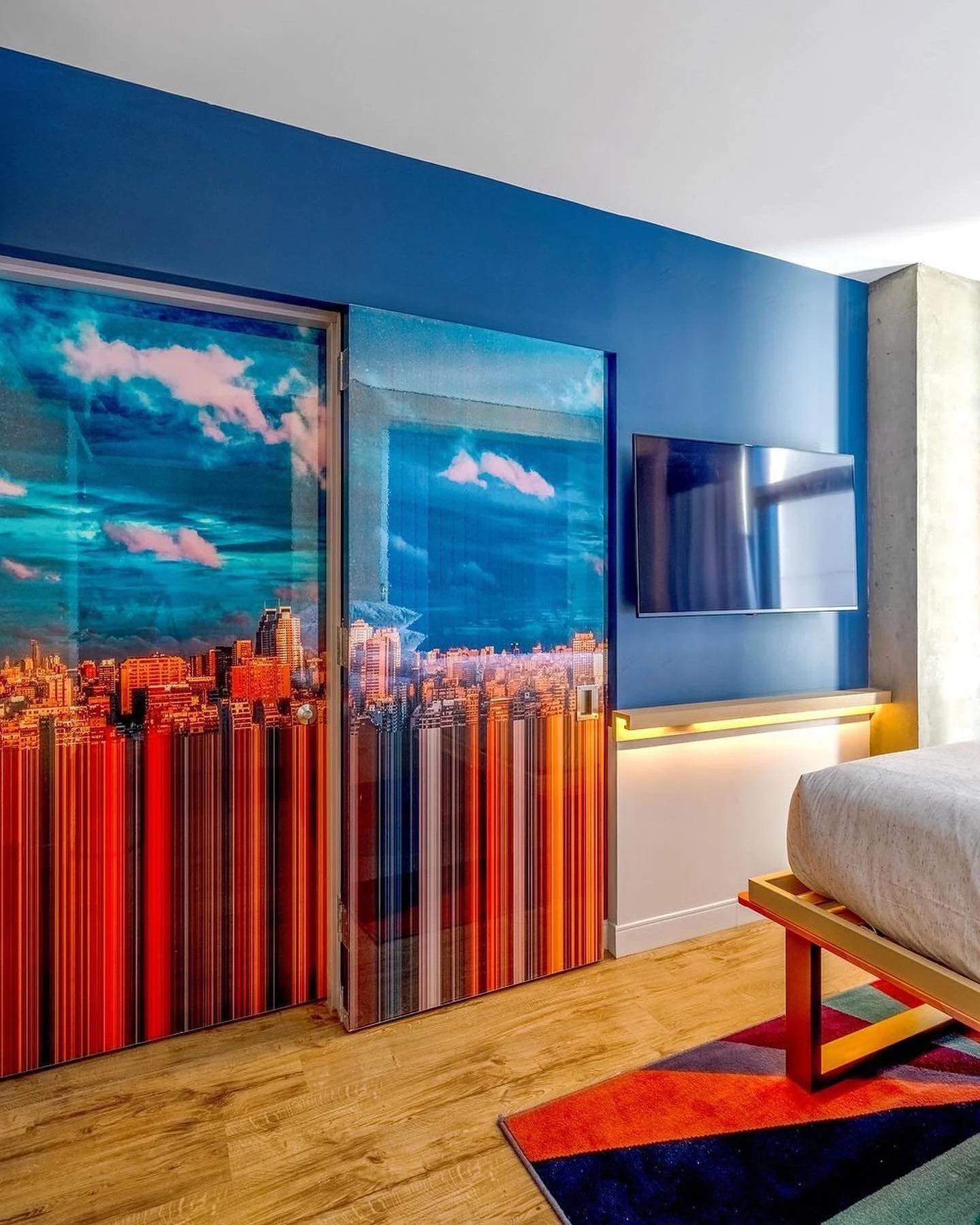 A bedroom with a blue wall, a flat-screen TV, and a colorful cityscape mural or wallpaper.