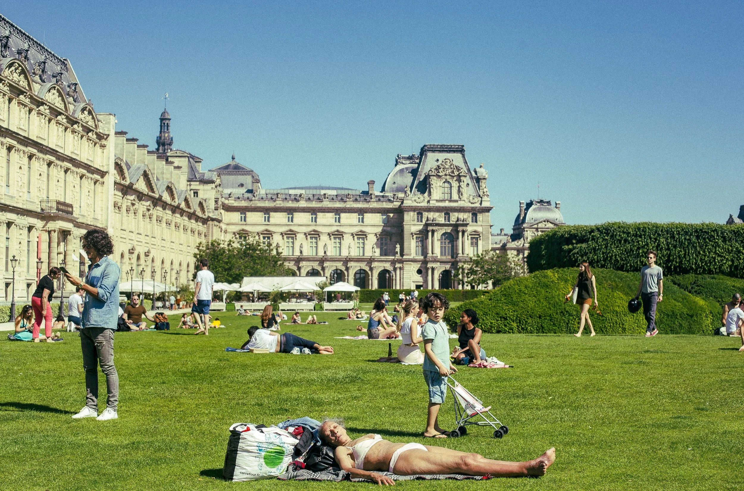 People relaxing and socializing on a lush green lawn in front of historic European-style buildings under a clear blue sky on a sunny day. Some are sitting, lying down, or walking, with a child standing near a stroller and a person lying on a blanket.