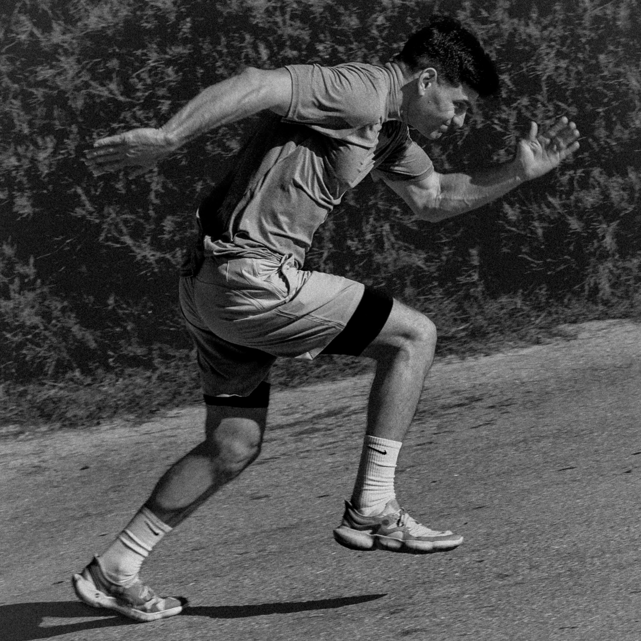 A young man is running on a track, wearing a T-shirt, shorts, and sports shoes. He is in a full sprint position, focused and determined, with trees and greenery in the background.