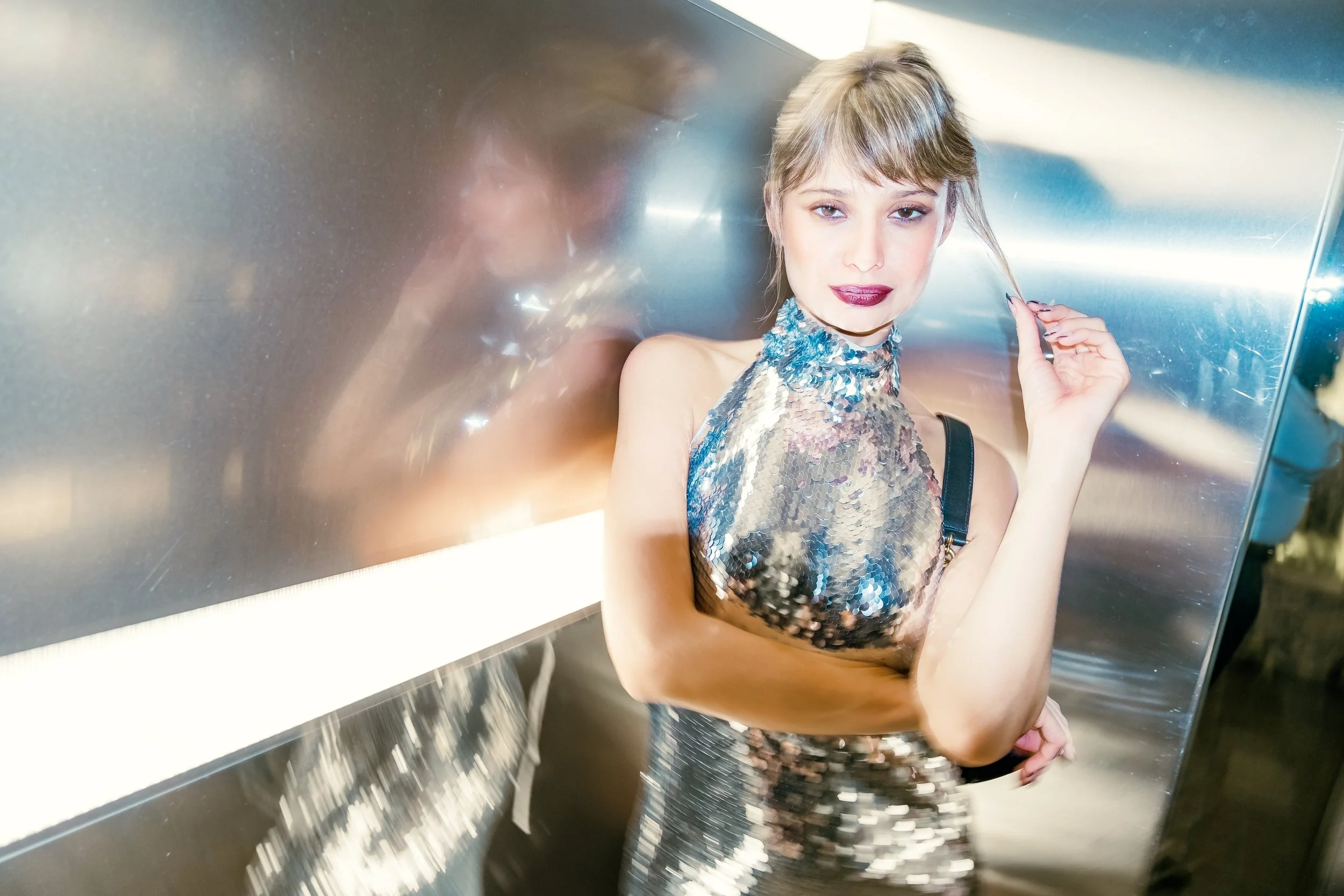 A woman with blond hair and makeup, wearing a shiny, sequined dress, standing in an elevator with metallic walls, looking at the camera.