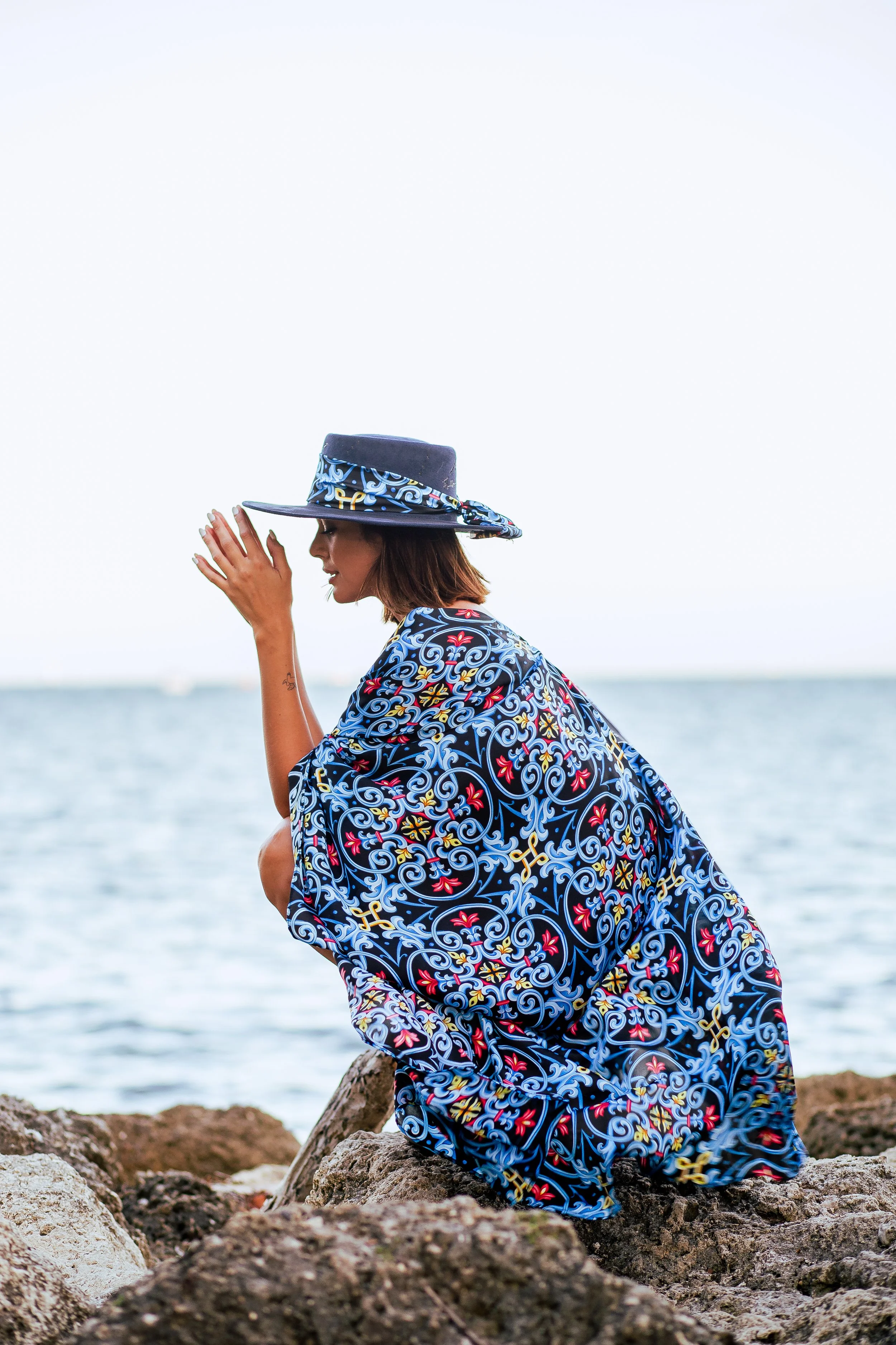 A woman wearing a colorful, patterned dress and a wide-brimmed hat with a matching design, sitting on rocks near a body of water, with a sky and horizon in the background.