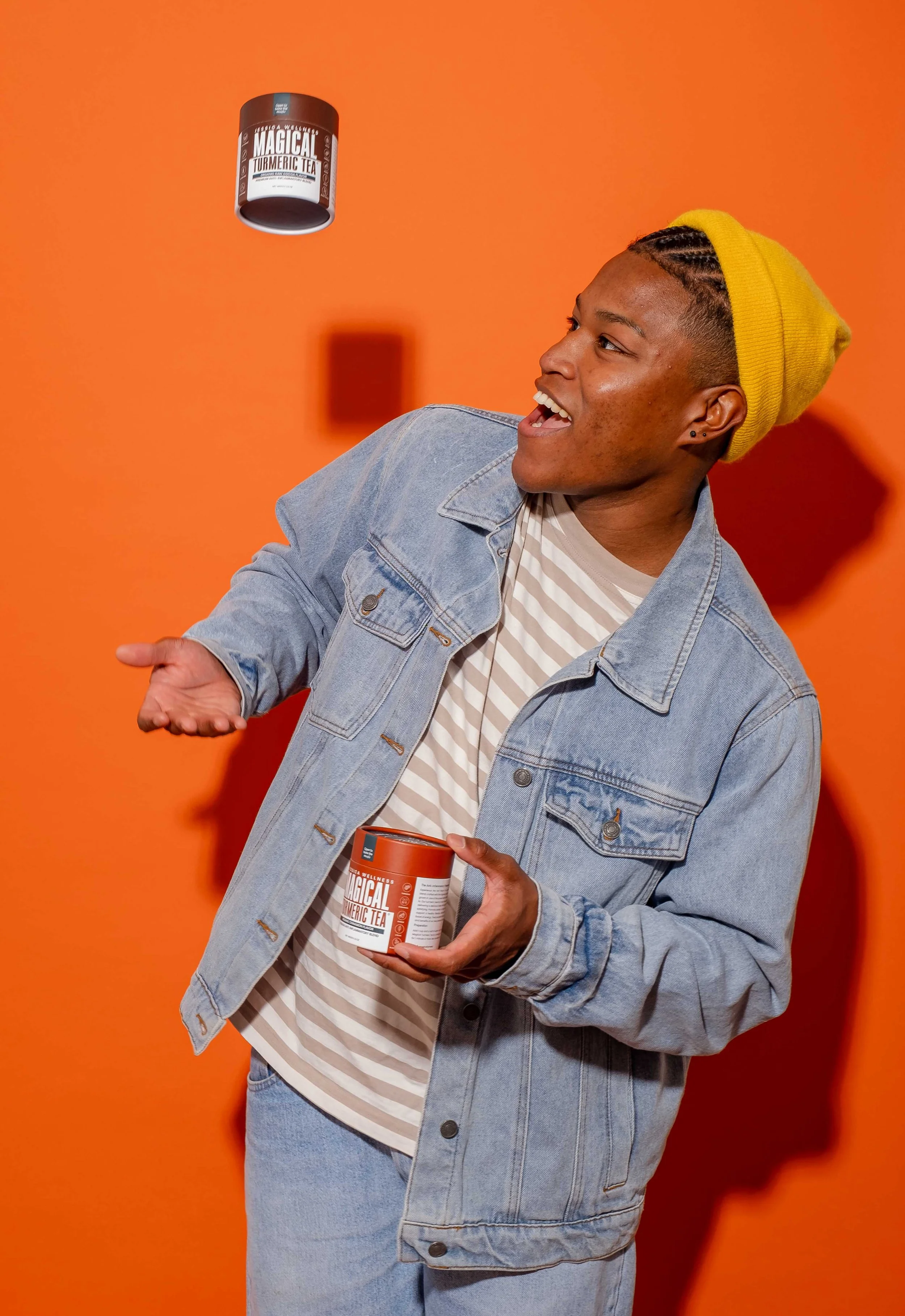 A person with a yellow beanie and denim jacket is holding a container of Magical Turmeric Tea while gesturing with one hand against an orange background.