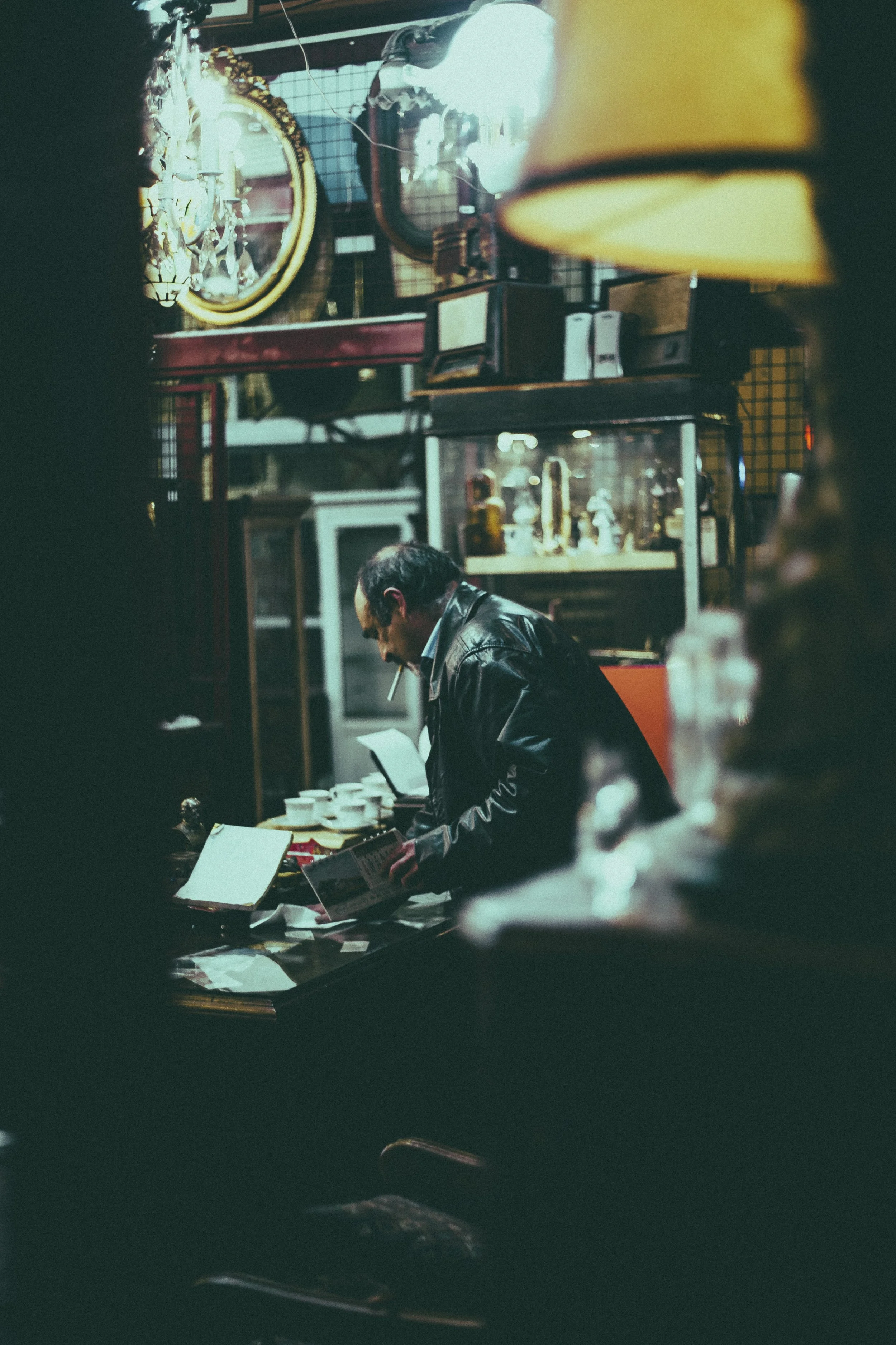 A man wearing a leather jacket is sitting at a table reading a newspaper, with various objects and a display case in the background, captured through a small opening framed by dark surroundings.