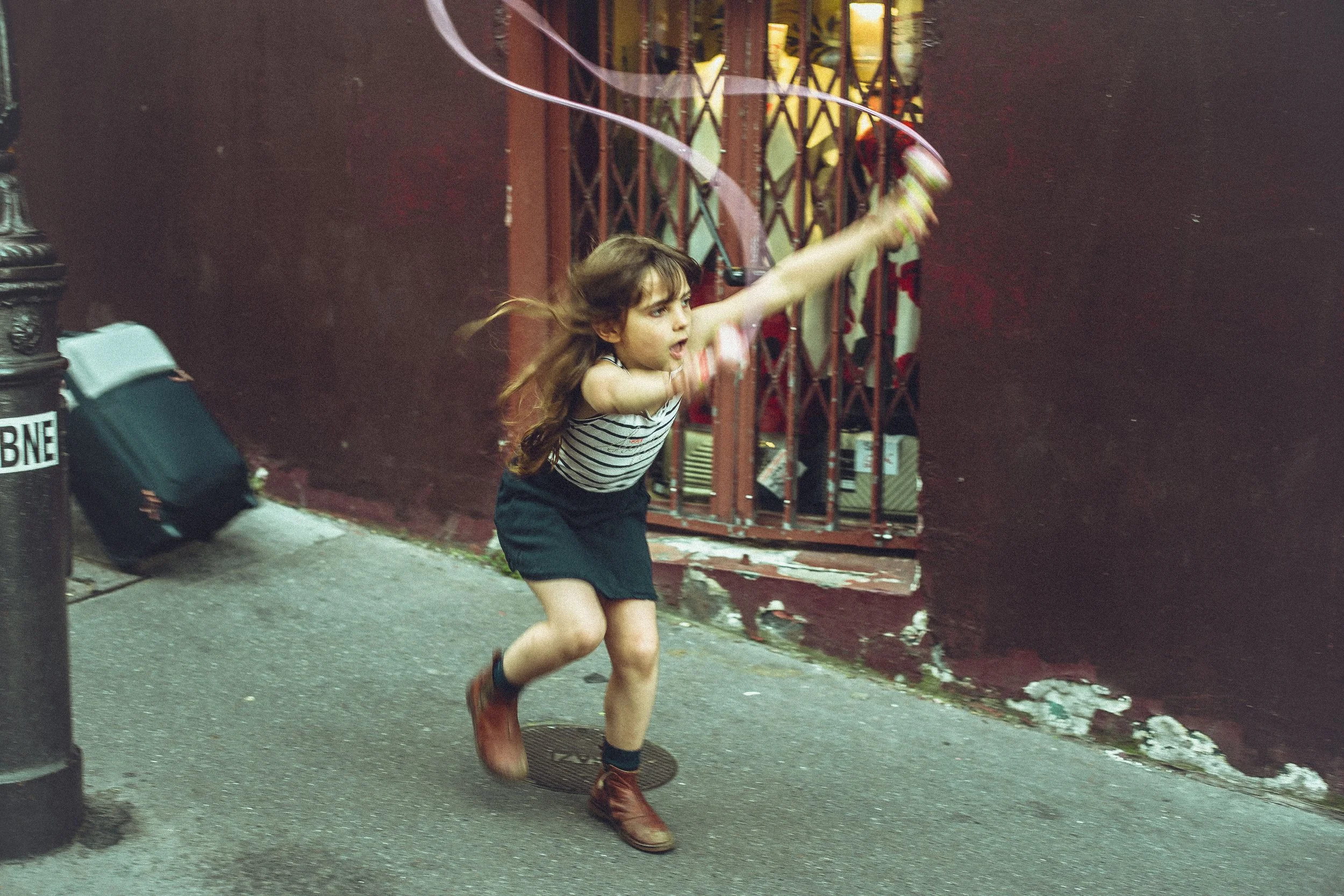 A young girl with long hair, wearing a striped tank top, shorts, and boots, is running and playing with a hula hoop outside next to a red gate and a trash can.