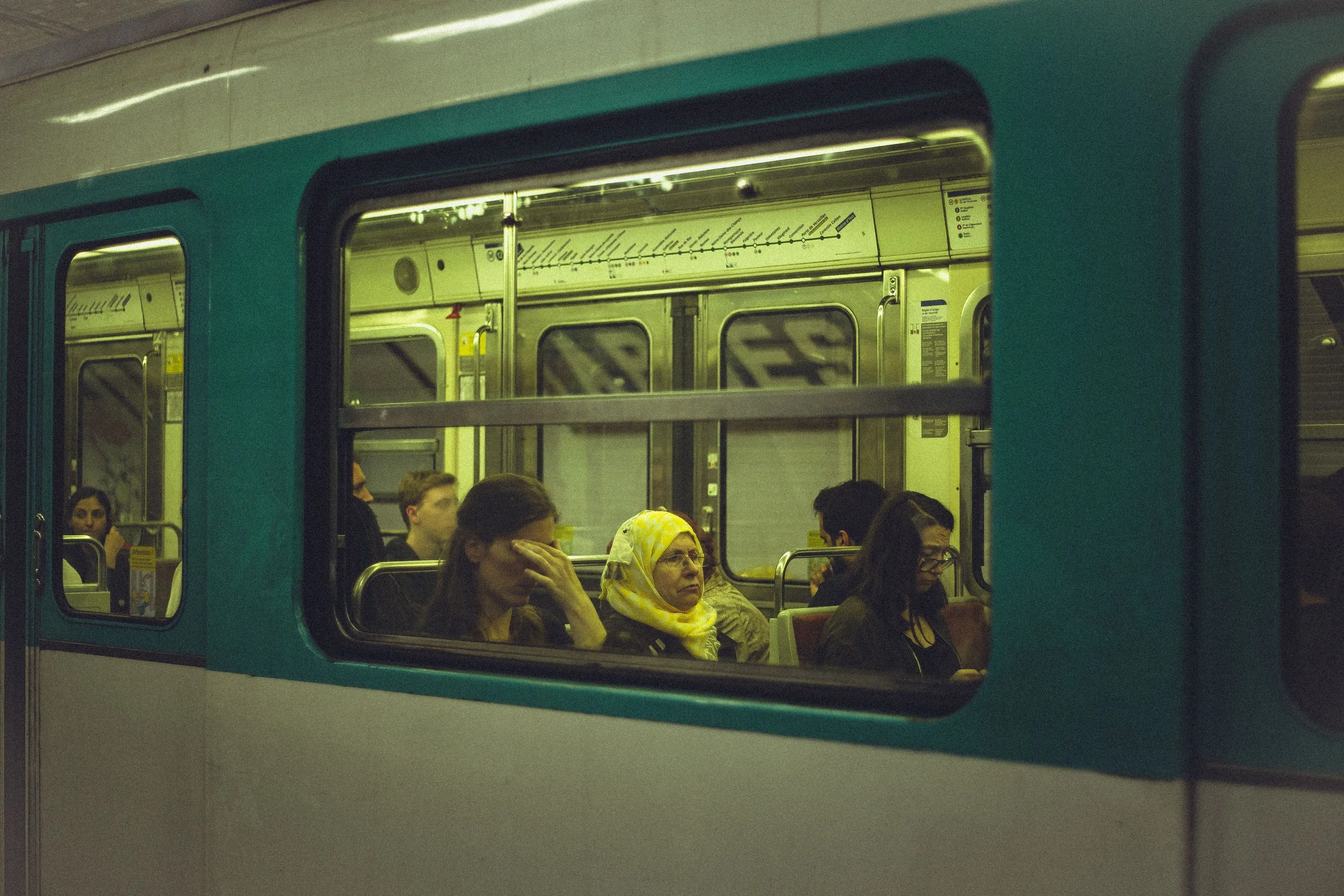 View through a subway train window showing passengers seated inside, including women and men engaged in various activities.