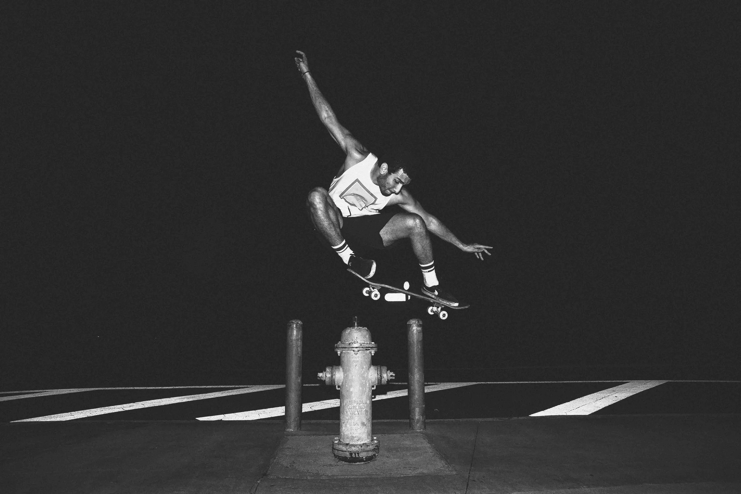 A man wearing a white sleeveless shirt and shorts skateboarding over a fire hydrant at night on a street.