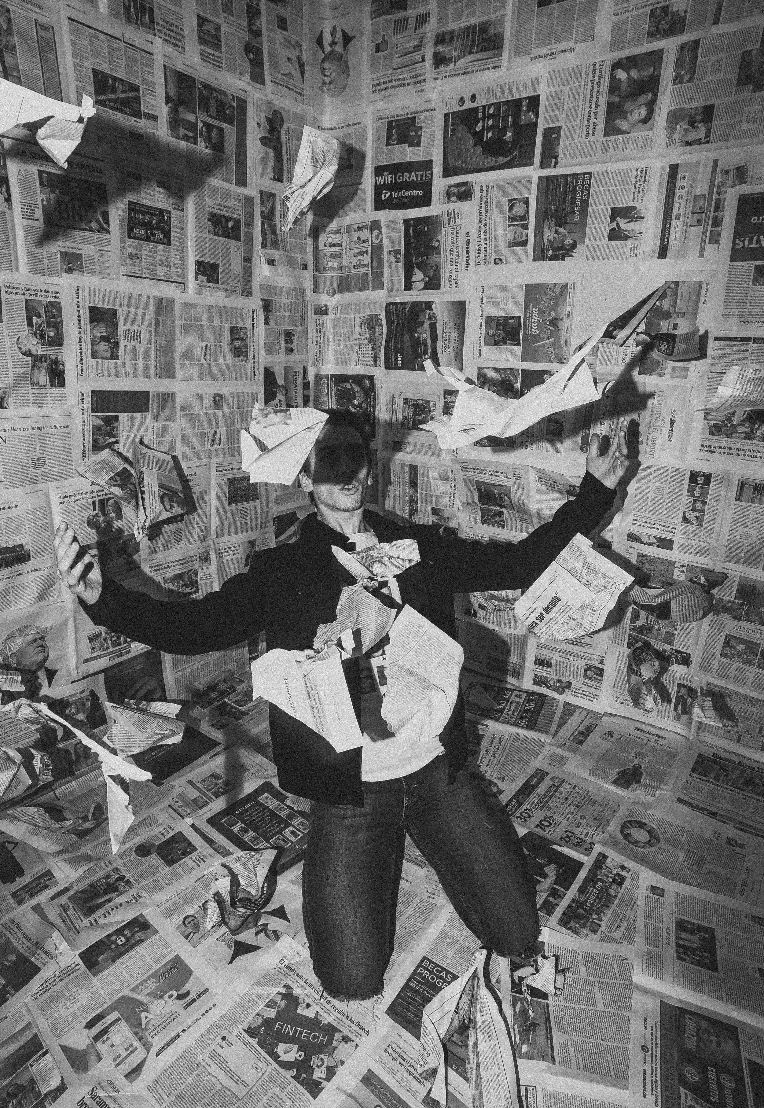 Person standing in a room with floor and walls covered in newspapers, holding some papers in both hands.